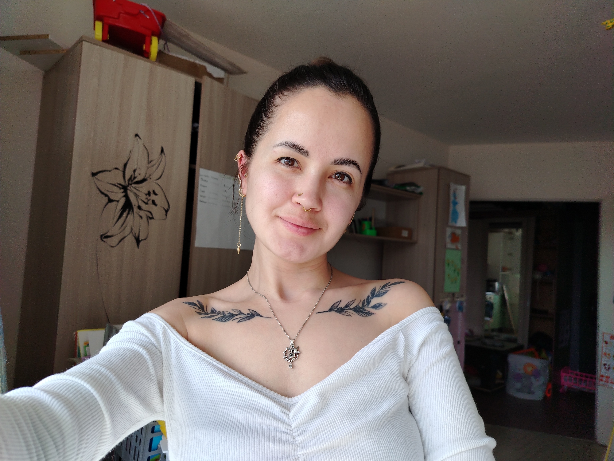 A young woman is centered in the frame, taking a selfie. She has dark hair pulled back in a bun, brown eyes, and light skin. She wears a white, ribbed, off-the-shoulder shirt. A silver necklace hangs around her neck, featuring a decorative pendant. She has two dark, leafy tattoos on her upper chest, just above her shoulders. Several earrings are visible in her left ear. She smiles slightly at the camera.
Behind her is a cluttered domestic interior. To her left, a light wood-grain wardrobe with a large stylized flower decal can be seen. A red toy box is on top of it. In the background is an open doorway that leads to another room with various items, including furniture and other toys, stored out of the light, indicating an unused area or storage space. The picture appears to have been taken in an apartment in the city of Noviy, Russia.
