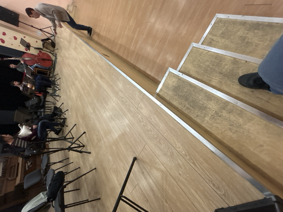 The image displays an indoor scene, identified as a musical rehearsal or performance space in Plovdiv, Bulgaria. The perspective is from the bottom of three light brown wooden steps, edged with silver metal, leading up to a raised stage. The photographer's feet, wearing black shoes and blue trousers, are visible on the lowest step.
On the light brown wooden stage, five male individuals are present. Four are seated, positioned with music stands and instruments. From the left, a man in a striped sweater holds a light-colored wind instrument resembling bagpipes. Adjacent to him, a man in a plaid shirt plays a stringed instrument, possibly a ukulele or mandolin. Further right, a man in a green top plays a red double bass. A fourth seated individual is partially visible further right. On the far right edge of the stage, a fifth man, wearing a light-colored jacket and dark trousers, is actively leaning forward with both arms extended, appearing to direct the musicians.
The stage is set against a dark background, revealing a large, antique-looking wooden structure resembling an organ on the far left. To the right, a white wall panel decorated with multiple red heart shapes is visible, next to a black speaker on a stand. Empty black chairs and additional music stands are arranged around the stage. The environment is illuminated by artificial lighting.