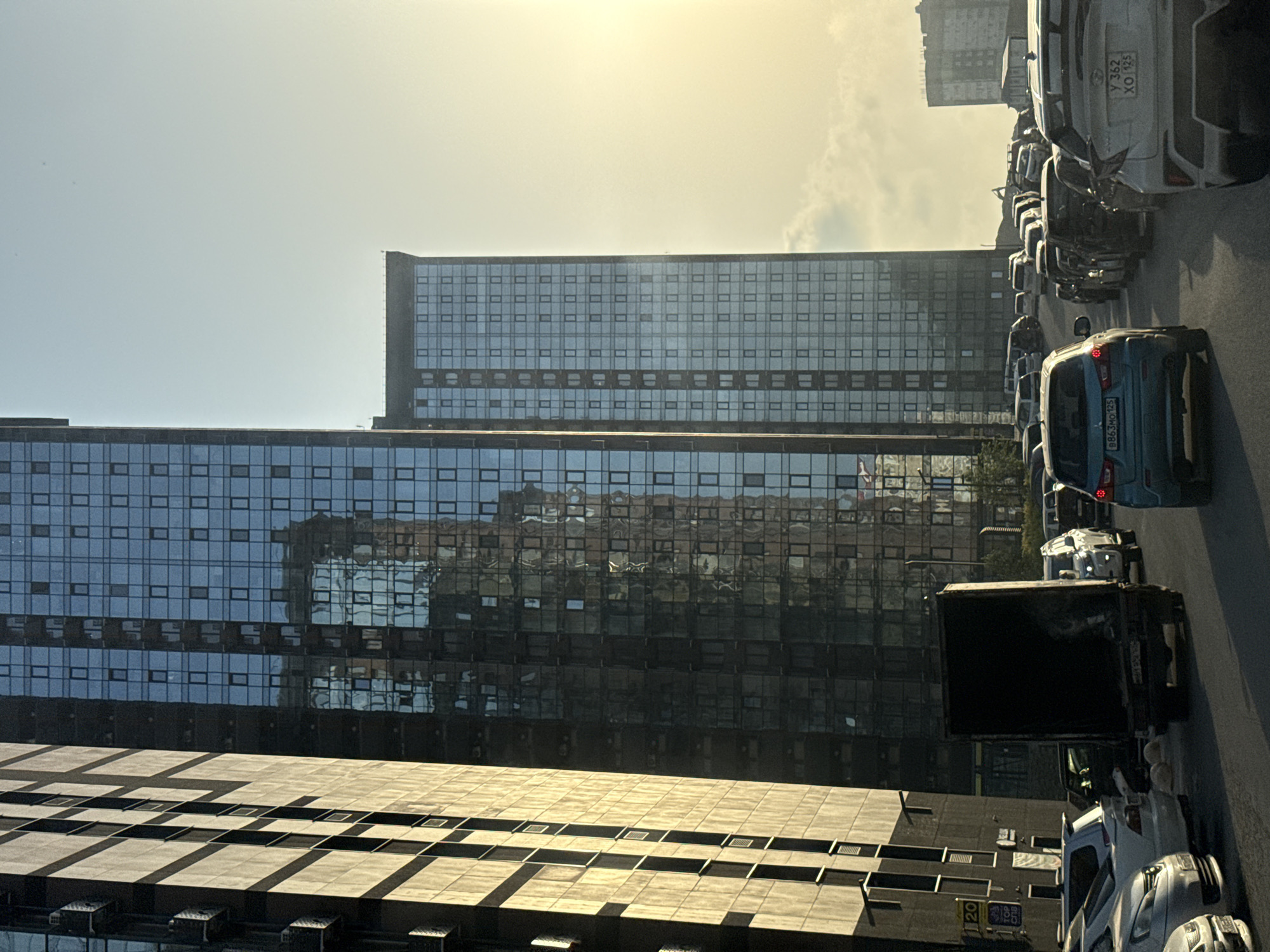 The image displays an urban street scene, focusing on modern architecture and parked vehicles under a bright, hazy sky. Three prominent buildings define the background: two tall, contemporary towers feature extensive glass facades that reflect the sky, clouds, and surrounding structures. In the foreground, a shorter, blockier building with multiple levels of rectangular panels and windows also captures sunlight, creating strong shadows.
The street, visible on the right side of the frame, is lined with numerous parked cars. A light blue SUV with the license plate "B863MO 125" is clearly visible, along with a white car bearing the plate "Y 362 XO 125". The "125" region code on both plates identifies the location as Primorsky Krai, confirming Vladivostok as the general area. Further down the street, a dark box truck with its rear cargo area open is parked. No individuals are clearly discernible.
The time of day appears to be either morning or late afternoon, indicated by the bright, somewhat low-angle sunlight creating stark highlights and shadows across the buildings and vehicles. The sky is mostly clear with a soft, warm haze and a few faint clouds. A small, partially obscured sign on the lower foreground building displays the number "20".