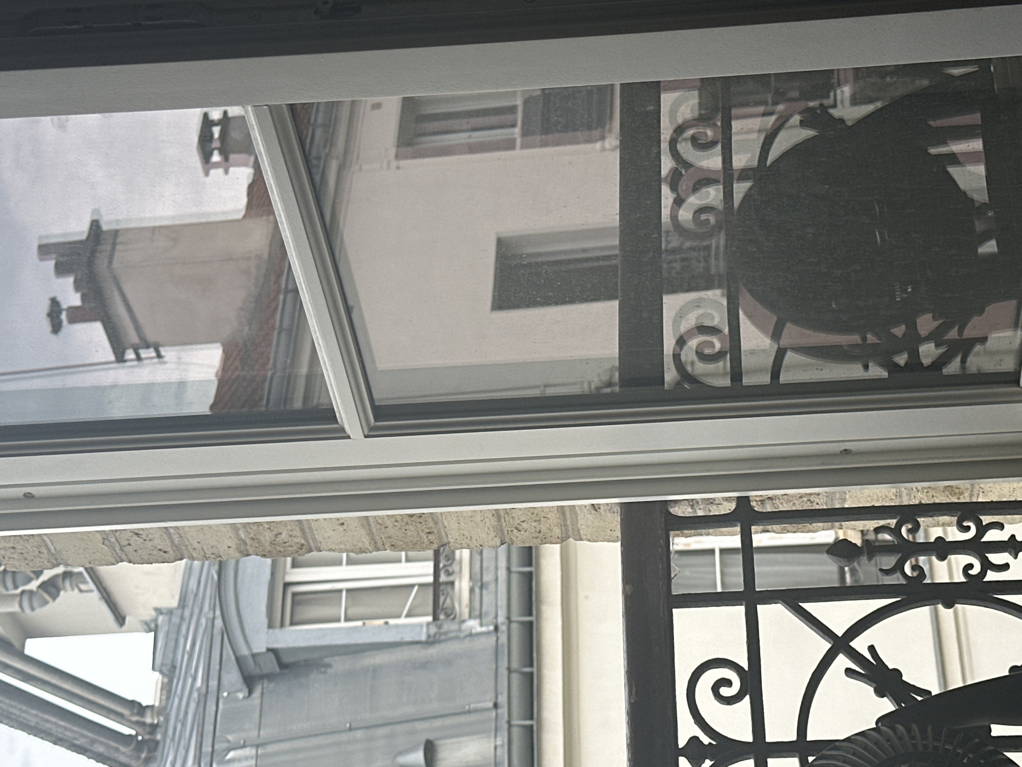 The image shows a view from a window looking out onto a Parisian street scene. In the foreground, there is a black wrought-iron balcony railing with ornate scrollwork. Through the window, we see buildings with traditional Haussmannian architecture, characterized by mansard roofs, dormer windows, and chimneys. The reflections in the glass reveal more of the interior of the room from which the photo was taken, including a dark, shadowy object that appears to be furniture. The sky is overcast, contributing to a muted color palette of grays, whites, and muted earth tones.