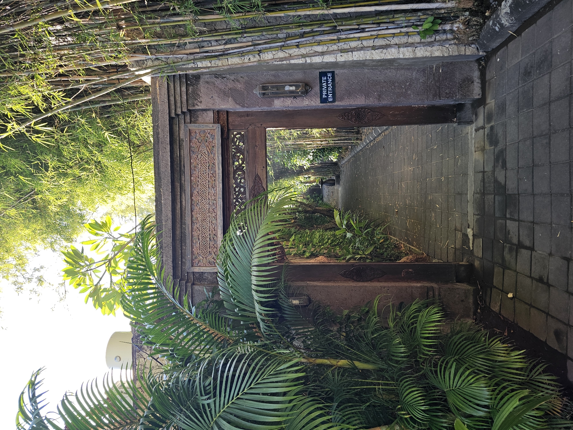 A pathway made of dark gray interlocking square pavers leads through a carved wooden archway. Lush green palm fronds frame the left side of the archway, while a dense grove of bamboo stands to the right. The archway is constructed of dark, weathered wood and stone, with intricate carvings above the opening. A small black sign with white lettering reading "PRIVATE ENTRANCE" is affixed to the stone pillar on the right side of the archway. A single light fixture with a frosted glass casing is mounted on the same pillar. The pathway continues into a verdant garden, suggesting a secluded or exclusive location. The lighting suggests it is daytime, with sunlight filtering through the trees. This appears to be the private entrance to a resort or upscale establishment in Denpasar, Indonesia.