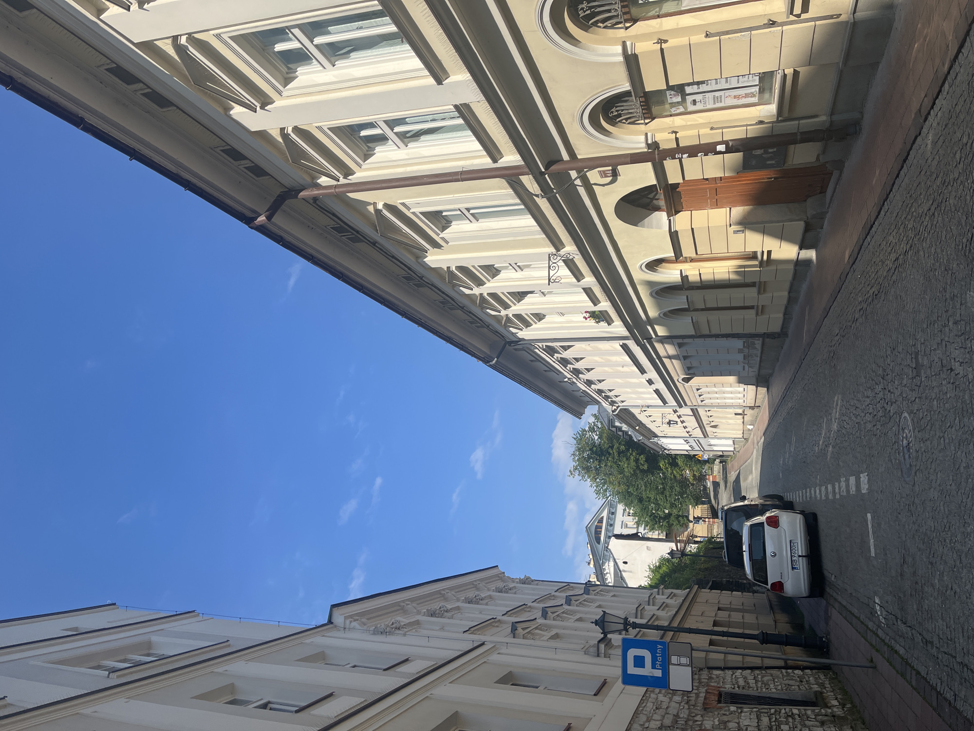 The image shows a street scene in a European city. The street is paved with cobblestones and lined with buildings on both sides. The buildings are old, with ornate facades and large windows. The street is relatively narrow and slopes uphill. The sky is blue with a few scattered clouds. There are two cars parked on the side of the street, and a blue parking sign with the word "Płatny" in white letters is visible on the left side of the street. A lamppost is also present near the parking sign. The overall impression is one of a historic European city center.