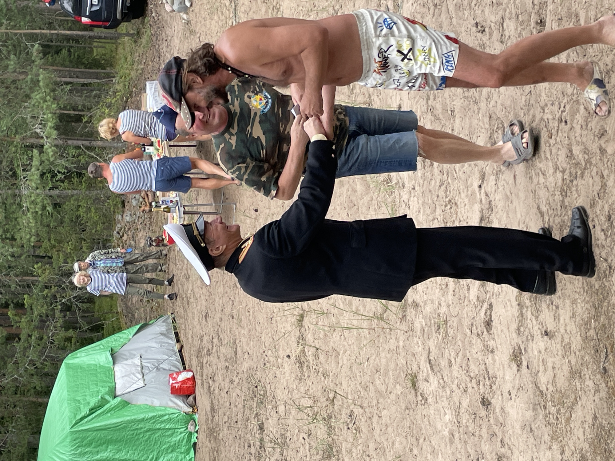 In Zelenaya Roshcha, Russia, a group of people are gathered on a sandy area surrounded by trees. In the foreground, a man in a dark uniform with a white hat is shaking hands with another man who is bare-chested and wearing patterned shorts. A third man, wearing a camouflage t-shirt and denim shorts, stands between them, also holding something in his hands. Further in the background, a green tent is set up, and several other individuals are visible. One person is standing near the tent, while others are near a table with food and drinks. A black SUV is parked in the distance. The ground is sandy with sparse vegetation.