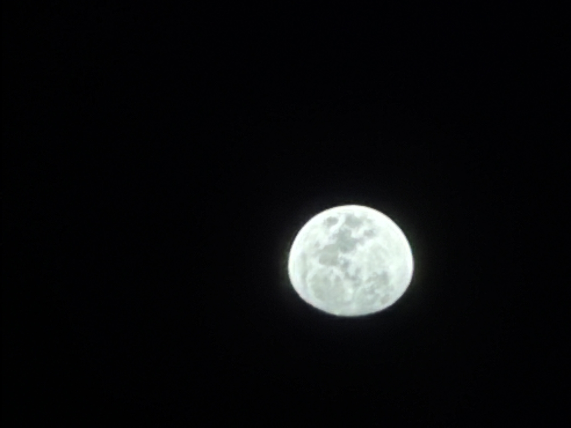 The media file is a photograph of a full moon in a dark, black sky. The moon is the sole subject, appearing as a bright, luminous orb with visible craters and surface details. It occupies the right side of the frame, with the majority of the image being the stark blackness of the night sky. There are no other objects or scenes visible, and no people are present. The image does not depict any specific activity or event beyond the presence of the moon. The time of day is clearly night due to the darkness and the illumination of the moon. There are no weather cues, and no location context is provided. The image is devoid of any text.