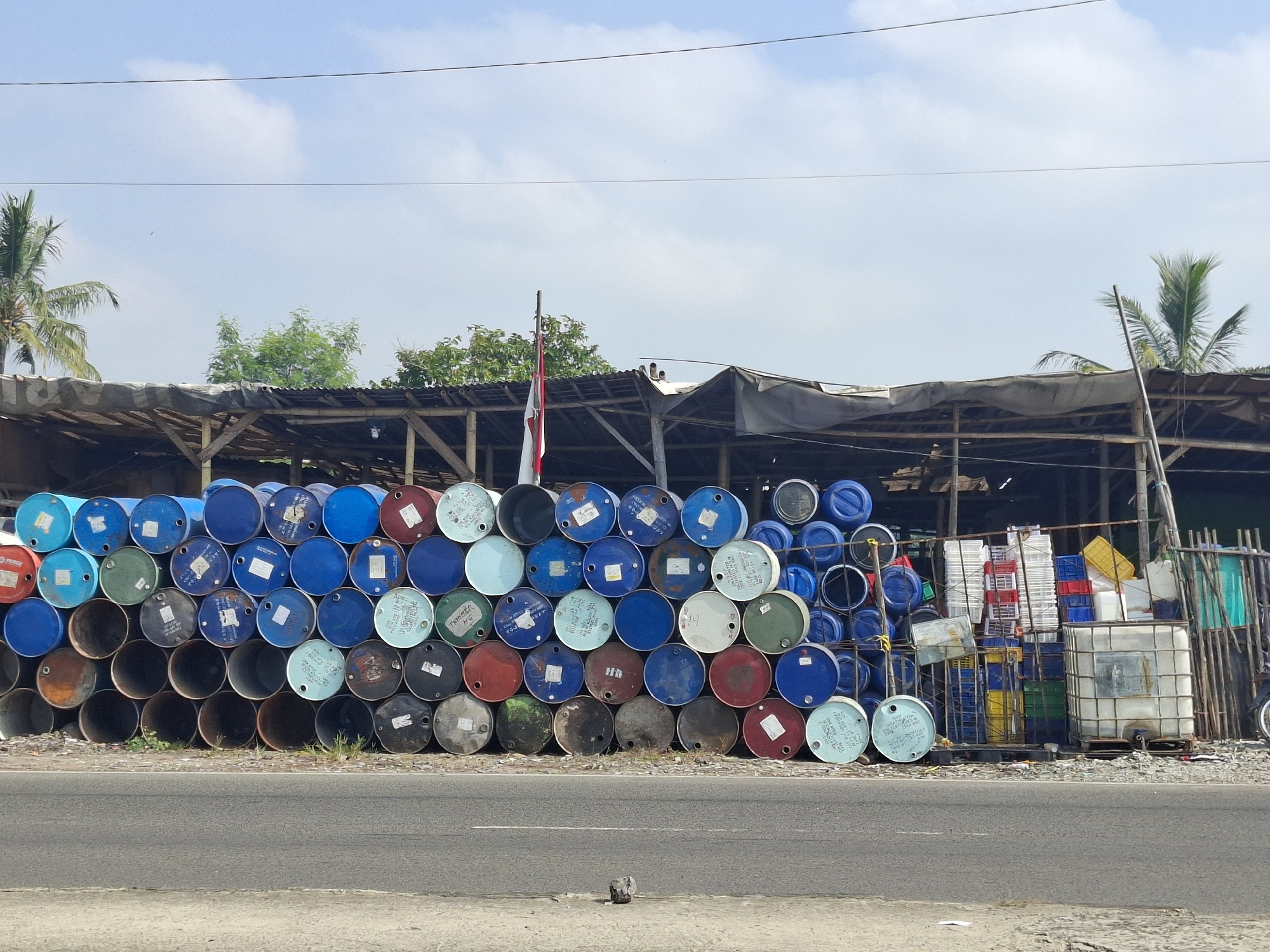 The image captures a scene in Sukabumi, Indonesia, featuring a large outdoor storage area filled with numerous cylindrical drums. These drums, of varying colors including blue, green, red, and white, are stacked high, forming a substantial wall. Behind the drums, a rustic open-sided structure, possibly a warehouse or storage shed, is visible with a corrugated metal roof supported by wooden beams. A flagpole with an Indonesian flag is positioned centrally behind the drums. Further to the right, additional storage containers, including white crates and blue plastic barrels, are visible. The foreground shows a paved road and a concrete curb. The environment appears to be an industrial or storage yard under a bright, partly cloudy sky, with palm trees visible in the background.