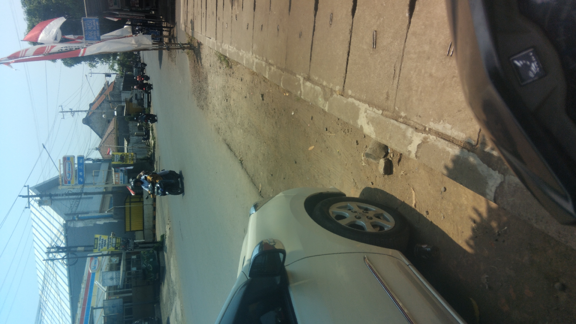 In a city street scene in Karangasem Selatan, Indonesia, the foreground is occupied by the partial view of a white car on the left and a dark motorcycle dashboard on the right. The road, paved with asphalt, is visible between the car and a concrete sidewalk. A person is riding a motorcycle on the road in the mid-ground, heading away from the viewer and towards the right side of the image. The background features buildings, including a structure with a silver, corrugated roof and an Indomaret store sign. There are also several Indonesian flags, some of which are red and white, displayed on poles. The environment appears to be a sunny day with clear skies.