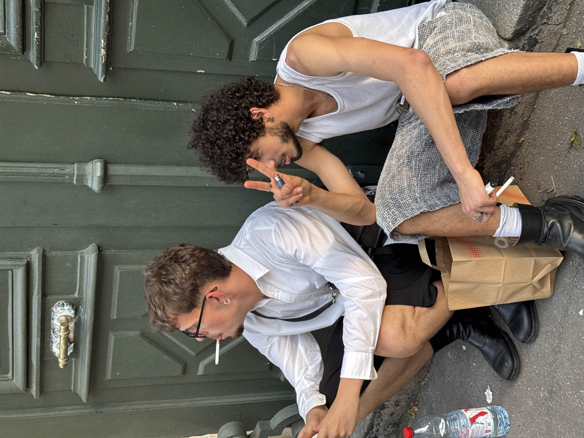 Two men are squatting outside a green, paneled door in Paris, France. One man, with curly hair and wearing a white tank top and patterned shorts, is holding up two fingers in a peace sign while holding a lighter. The other man, with glasses and a white button-down shirt, is smoking a cigarette. There is a brown paper bag and a water bottle on the ground beside them. Both men are wearing black boots.