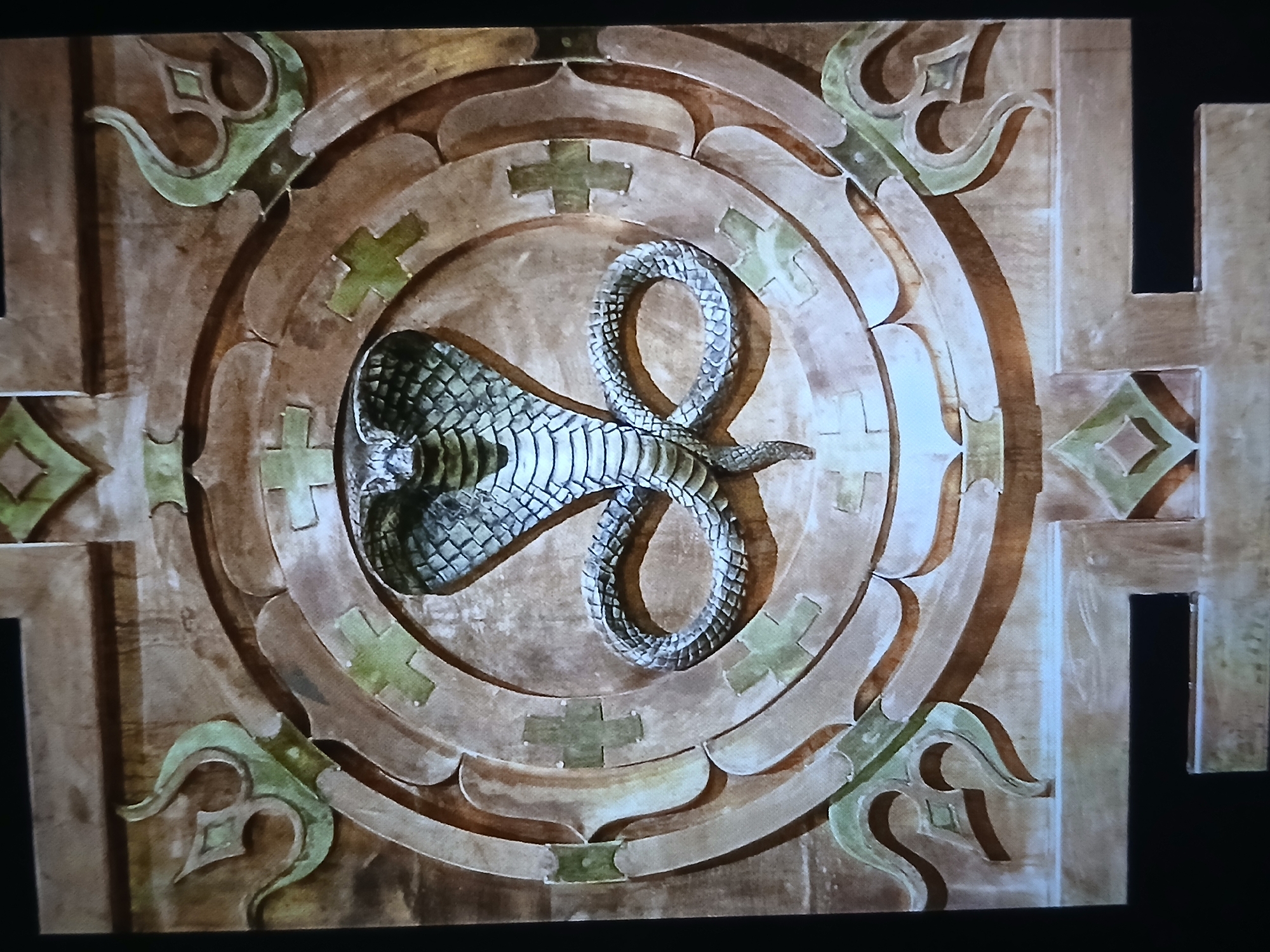A close-up shot shows an ornate circular carving with a snake motif. The carving appears to be made of wood or stone, with a brown and beige color scheme. A cobra is centrally placed in the circular design, with its head raised and its body coiled into an "S" shape. The snake's scales are detailed, and it looks almost metallic. The outer edges of the circular carving feature decorative elements that include stylized crosses and other geometric patterns in green. The entire carving appears to be part of a larger architectural feature. Given the likely location being Bengaluru, India, it is possible that this carving is part of a temple or a building with Hindu or Southeast Asian artistic influences. There is no presence of people in this image.