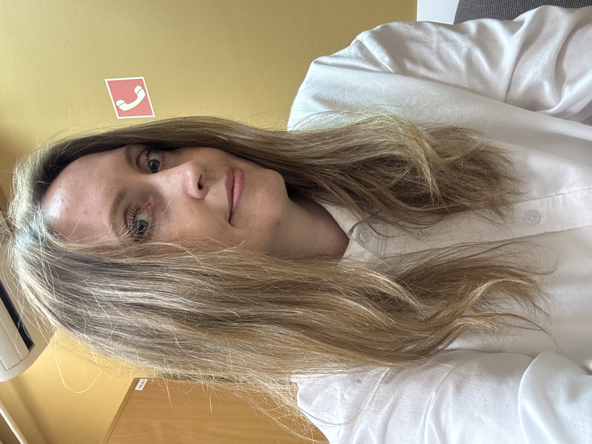 An indoor, close-up shot features a woman with long, wavy, light brown and blonde hair, looking directly towards the camera with a calm, subtle smile. She has fair skin and is wearing a white collared shirt. Her hair frames her face and cascades over her shoulders and chest.
The background is dominated by a plain yellow wall. Prominently displayed on this wall, above and slightly to the left of the woman's head, is a rectangular red sticker with a white telephone receiver icon, signaling a communication point. To the far left of the image, a white object, possibly part of an air conditioning unit or vent, is visible. A light brown wooden surface, likely a desk or table, is partially seen in the lower left corner. To the right, behind the woman's shoulder, a textured grey surface, possibly upholstery from a chair or furniture, completes the background.
The lighting is bright and even, suggesting an indoor setting during the daytime or a well-lit interior. The woman's expression is relaxed and pleasant, indicating a comfortable pose for the photograph. The scene suggests an office or public building in Chaikovsky, Russia. There is no discernible text beyond the stylized phone icon.