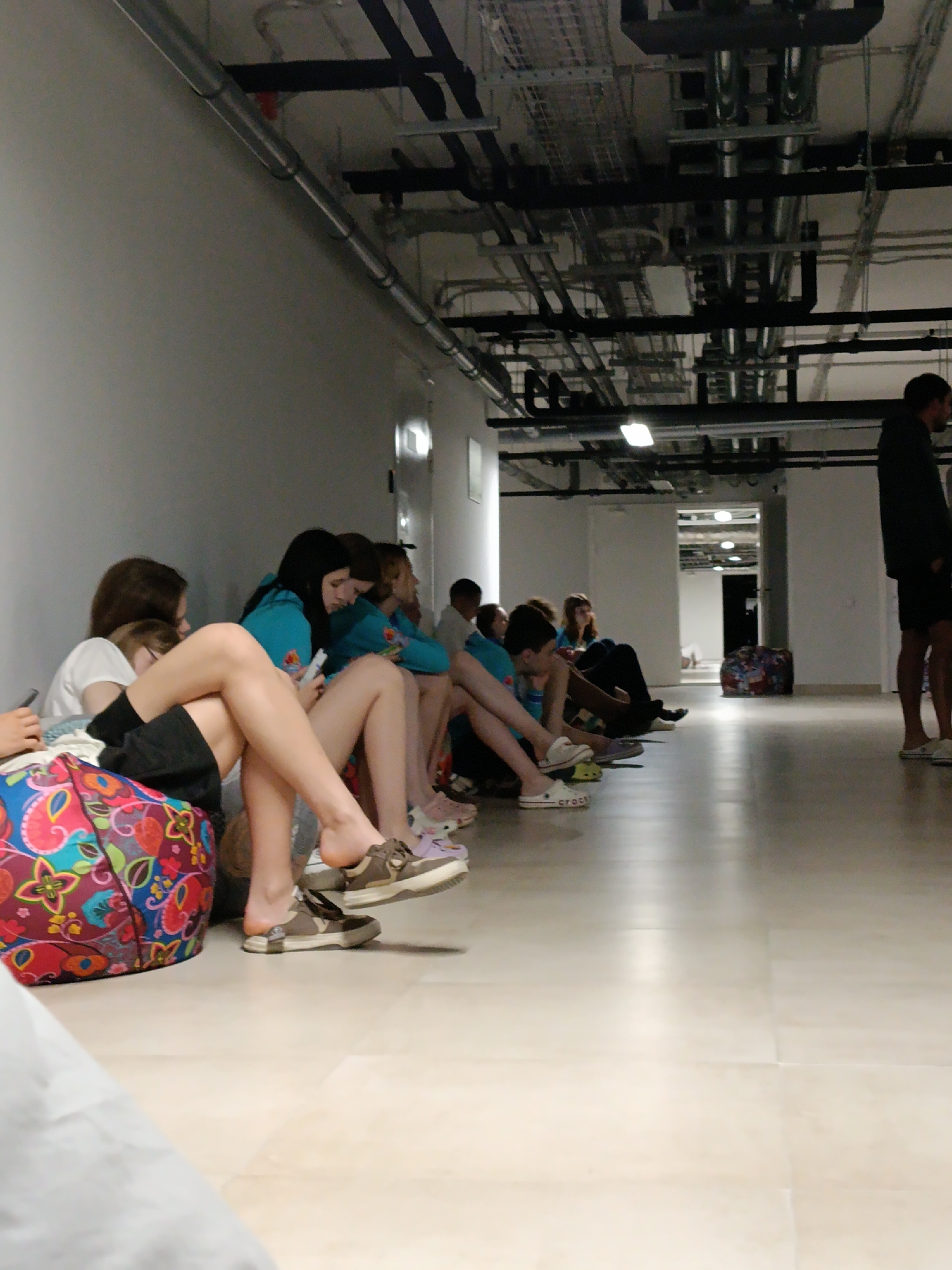 The image captures an indoor scene within a long, utilitarian-style corridor or hallway. A group of young people, mostly females, are seated on the light-colored tiled floor along a white wall.
In the foreground and mid-ground, several individuals are visible, many casually dressed in t-shirts, shorts, and hoodies. Some are observed looking at their mobile phones, suggesting a period of waiting or leisure. One person's bare feet and legs are prominent in the foreground, with one foot partially on a shoe where the brand "crocs" is visible. Other footwear includes sneakers and sandals. A vibrant, multi-colored beanbag or pouf with a floral and paisley pattern is positioned next to a seated individual on the left.
The ceiling features exposed industrial-style infrastructure, including silver pipes, black ducts, and various lighting fixtures, indicating a modern or renovated building. Further down the corridor on the right, an adult male stands facing away from the camera. In the distance, another doorway opens into a dimly lit space. The overall impression is a relaxed, informal setting, possibly a waiting area in a school, convention center, or similar public building. The lighting is artificial, illuminating the hallway uniformly. No other explicit text is visible.