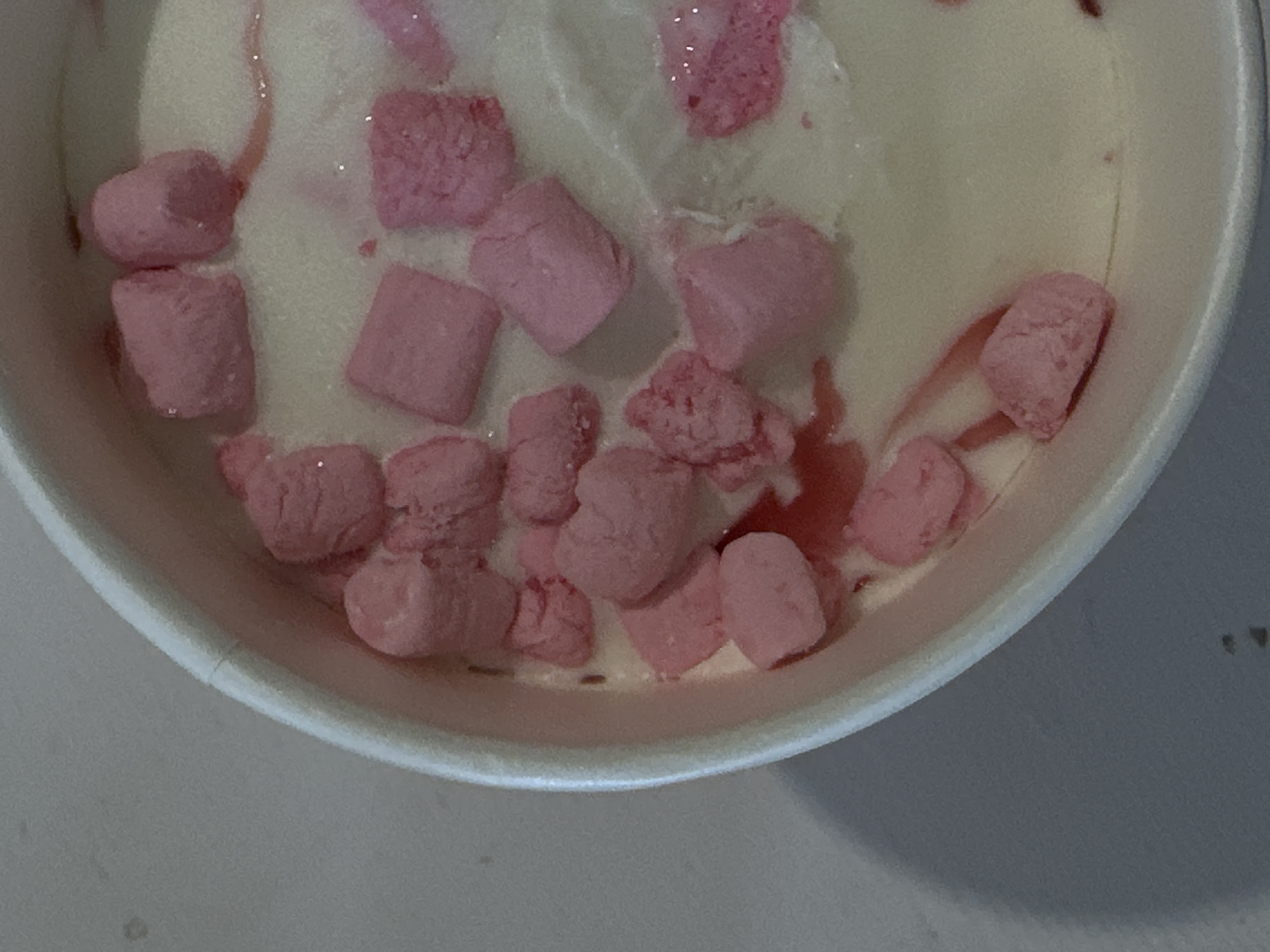 A close-up, overhead shot shows a partially consumed serving of ice cream in a white paper container. The ice cream is light-colored with irregularly shaped pink marshmallow pieces distributed throughout. Traces of a reddish-pink liquid are visible, likely syrup or melted marshmallow. The container's rim is visible and slightly out of focus. The background is a plain, light-colored surface. There is no evidence to suggest a location within the city of Votkinsk, Russia. There are no people or subjects of interest.