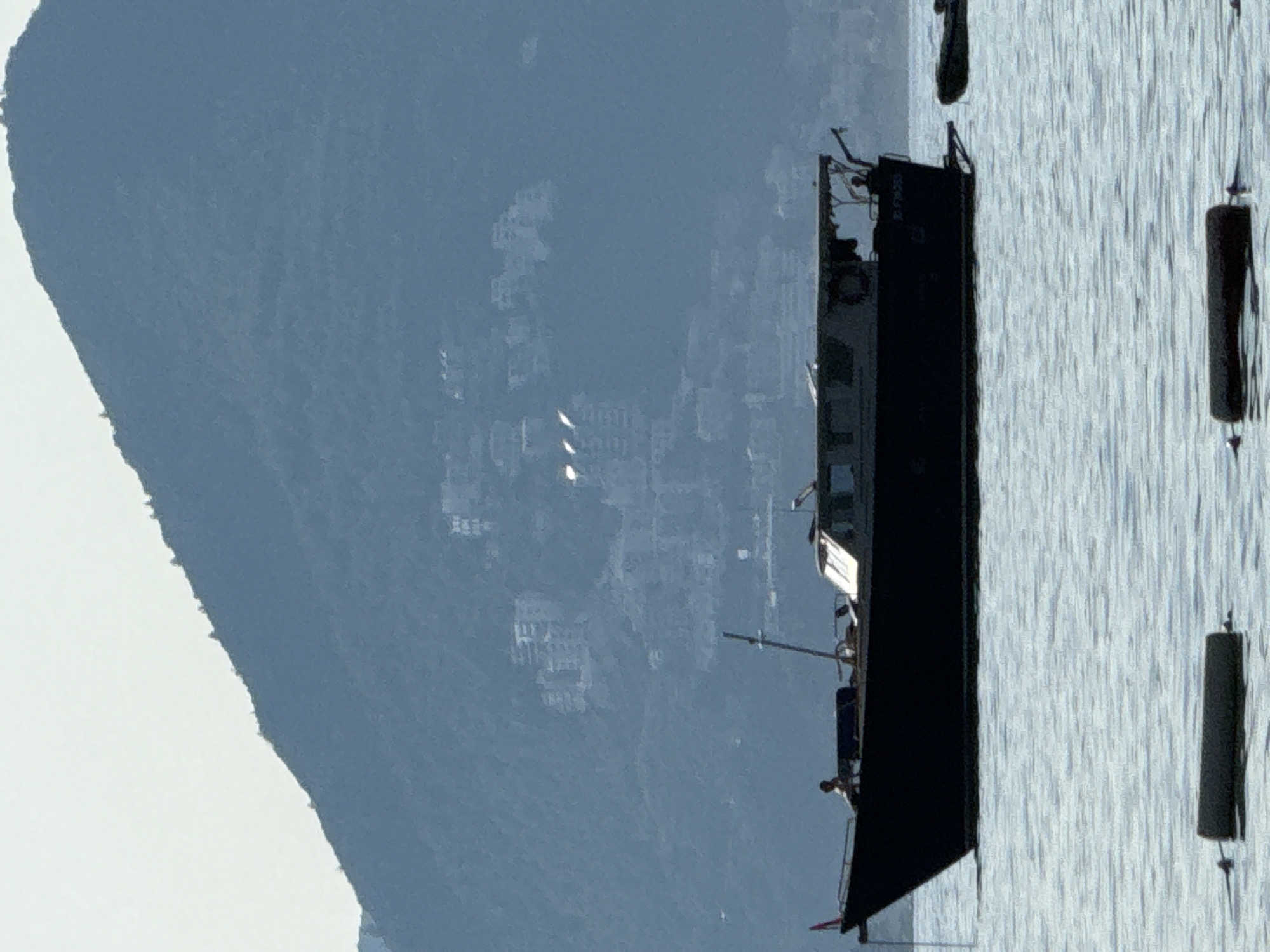 In Sveti Stefan, Montenegro, a dark-hulled motor yacht is anchored in calm, rippling water. The yacht features a cabin with a white roof and windows, and a mast rises from its deck. A person is visible on the foredeck, and another appears to be near the stern. In the background, a large, forested mountain dominates the scene, with scattered buildings visible on its slopes. Several cylindrical buoys are floating on the water in the foreground. The sky is a pale, hazy blue.