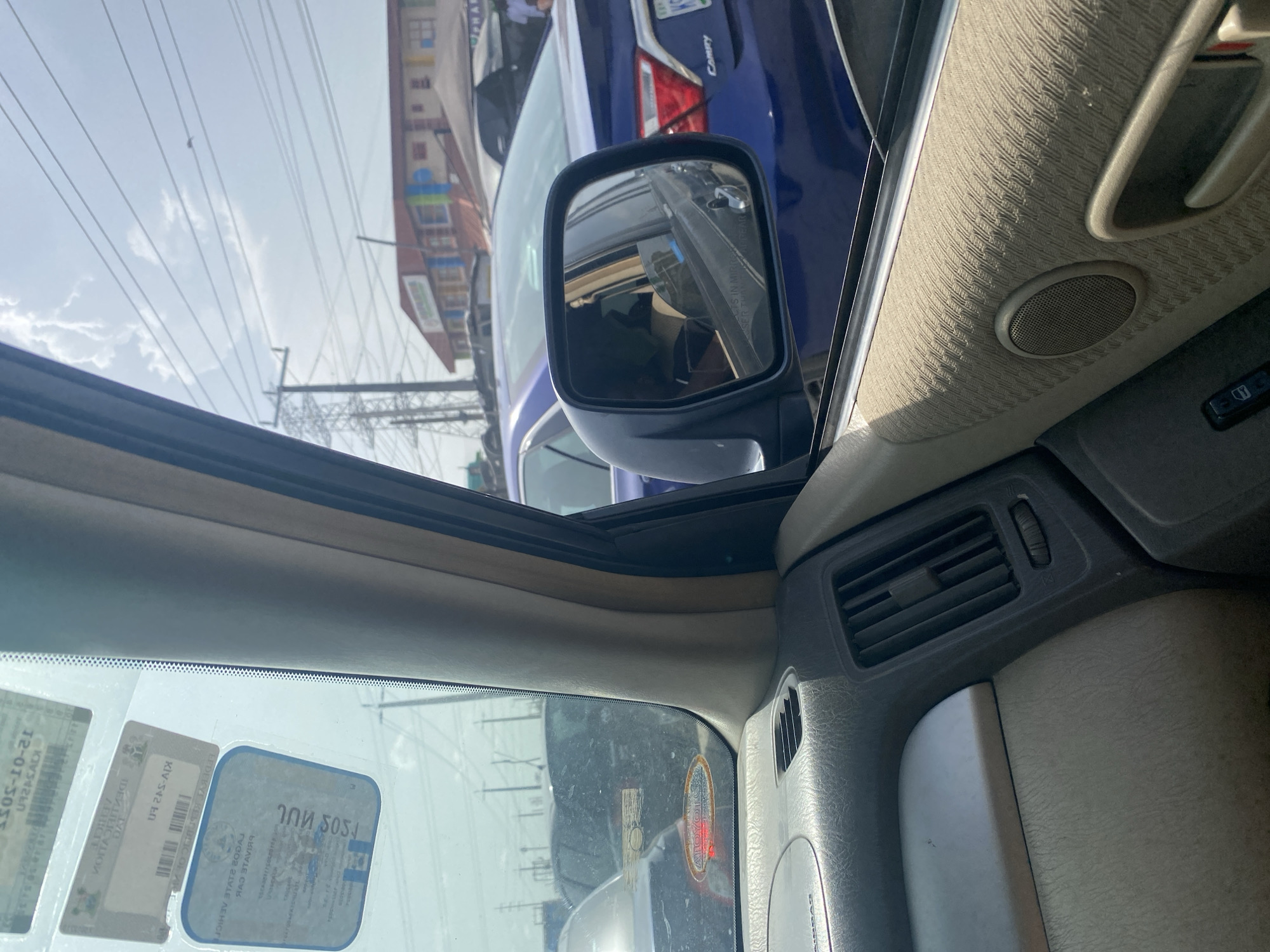 The image captures a view from inside a car, looking outwards through the passenger side window, with a significant portion of the car's interior visible.
**Subjects:** The interior of the car includes a dashboard, air vents, a speaker on the door panel, a fabric-covered pillar, and the side view mirror. The windshield features several official-looking stickers. Outside the car, power lines crisscross against a bright sky with scattered clouds. Several buildings are visible in the background. A blue sedan, identified as a "CAMRY" from its rear badge, is parked or driving alongside. A reflection of a person, likely the driver or a passenger, wearing a light-colored top, is visible in the side mirror.
**Activity or event taking place:** The car appears to be stationary, possibly in traffic or parked on a street, given the presence of other vehicles and roadside buildings.
**Notable details:** The scene is set during bright daylight hours, indicated by the clear sky and strong lighting. The presence of power lines and multi-story buildings suggests an urban or suburban environment. A key location cue is the text "LAGOS STATE GOVT PRIVATE CAR" visible on one of the windshield stickers, strongly indicating the location as Lagos State, Nigeria.
**Visible text:**
- On the blue car's rear: "CAMRY", and part of a license plate that includes "KIV-542 EN".
- Reflected in the side mirror: "OBJECTS IN MIRROR ARE CLOSER THAN THEY APPEAR".
- On windshield stickers (reversed due to reflection, corrected by OCR): "12-01-305", "LAGOS STATE GOVT PRIVATE CAR", and a series of numbers and letters like "7ՈՈ 5051", "HICK", "VOITAS", "DAT", "TSCAN", and a barcode.
