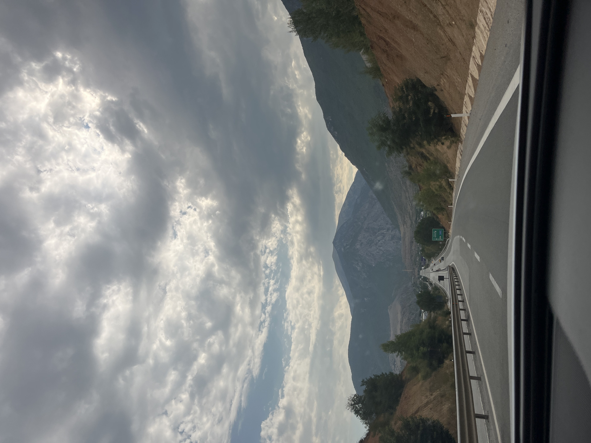 The image shows a view from inside a vehicle, looking out onto a highway in a mountainous region. The highway curves to the left, with a metal guardrail along the right side. The road is paved, with white lane markings. To the right of the road, there is a steep embankment with reddish-brown soil and sparse vegetation. Further up the embankment, there are trees.

In the distance, the highway leads towards a valley with buildings and more mountains. The mountains in the background are tall and rugged, with some rocky outcrops. The sky is filled with dramatic, heavy clouds, with patches of lighter sky and sunlight breaking through. The overall impression is of a vast, natural landscape with a man-made road cutting through it. There is a green sign visible in the distance on the right side of the highway, indicating a direction or destination. The time of day appears to be late afternoon or early evening, given the lighting and cloud cover.

The image appears to be a view of a highway leading towards the city of Bashkia Kukës, Albania, given the visual cues of mountainous terrain and road infrastructure consistent with that region.