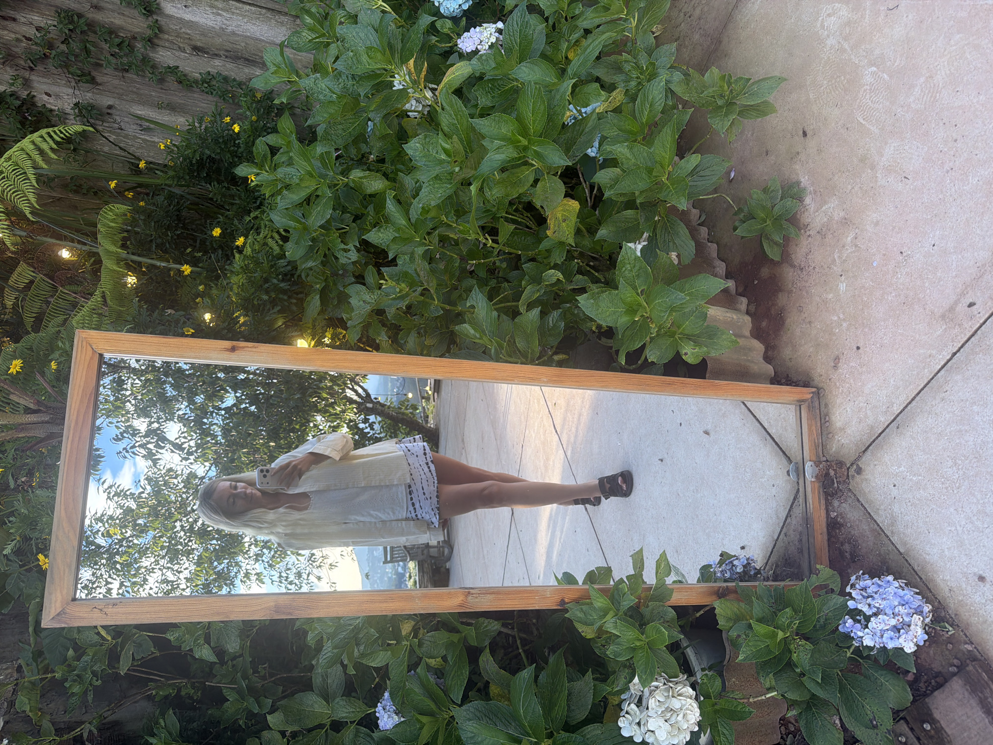 A young woman stands in front of a large, free-standing mirror. The mirror reflects the woman, who is wearing a light-colored outfit and holding a smartphone. She appears to be taking a selfie. The mirror is positioned outdoors, in what appears to be a garden. Lush green foliage and flowering plants surround the mirror, and a paved walkway is visible in the foreground. The background of the reflection shows trees and a bright sky, suggesting it is daytime. The scene is set in Xuân Thành, Vietnam.