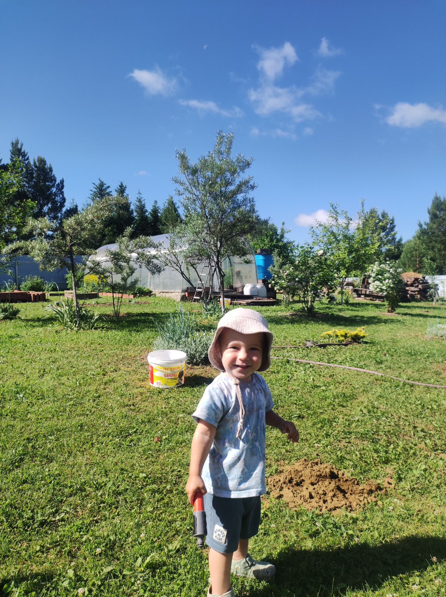 A full, slightly low-angle shot captures a young child in a sun hat and t-shirt standing in a grassy yard on a sunny day. The child is smiling at the camera, holding a red-tipped gardening tool. Behind the child, a bucket with yellow and red labeling sits on the grass. In the background, a greenhouse, trees, bushes, and a stack of lumber are visible, suggesting a rural or suburban setting. The sky above is a clear blue with a few white clouds. The location is identified as Fokino, Russia.