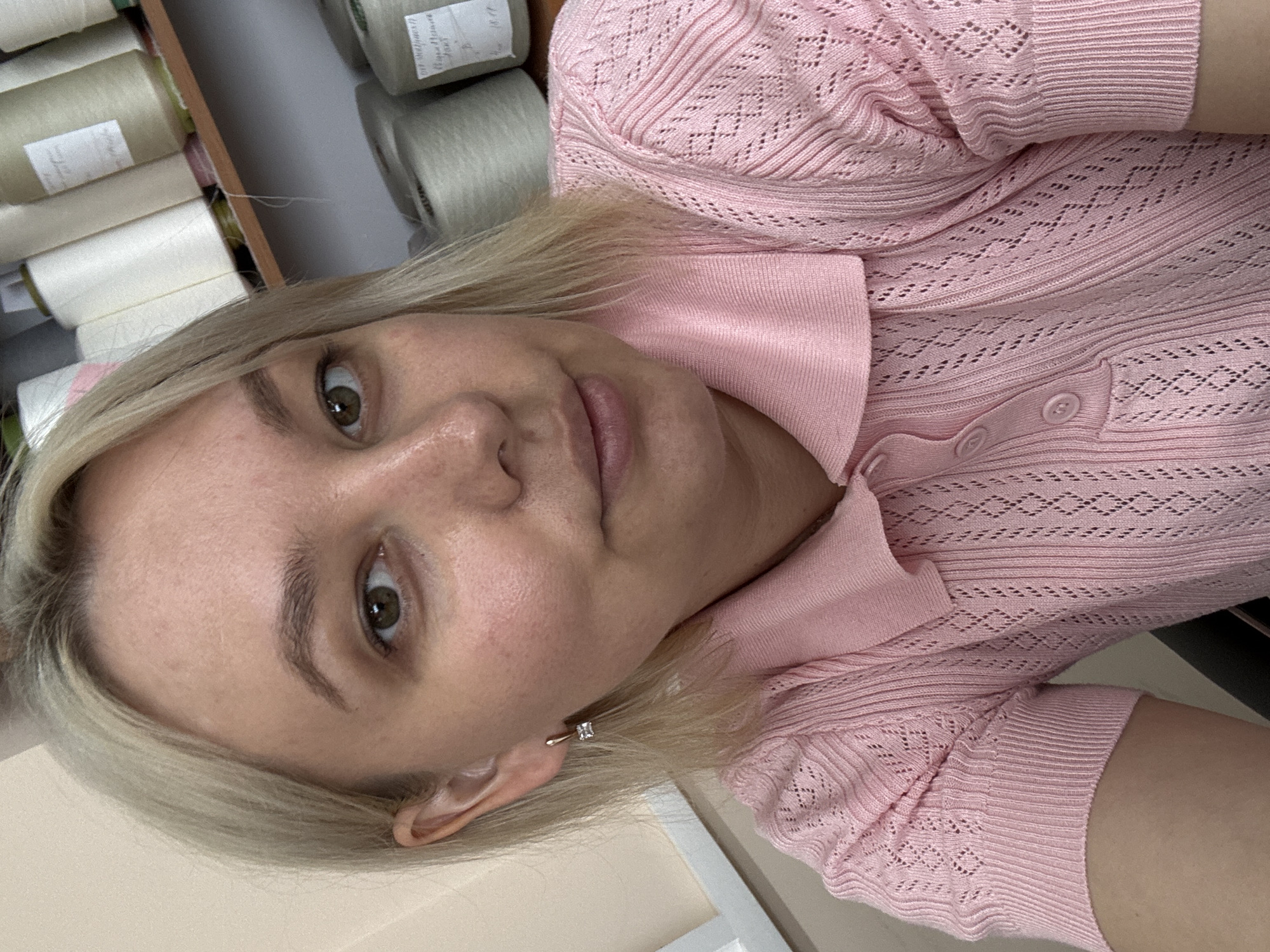 A medium shot shows a Caucasian woman with blonde hair and light skin facing the camera. She is wearing a pink knit polo shirt with a collared neckline and short sleeves. The shirt has a patterned texture with open diamond shapes. She is also wearing a small square earring on her left ear. The background is blurred, but appears to be a shelf filled with spools of thread, suggesting a fabric or sewing-related environment. There are no discernible landmarks or features that would indicate a specific location within the city of Chaikovsky, Russia.
