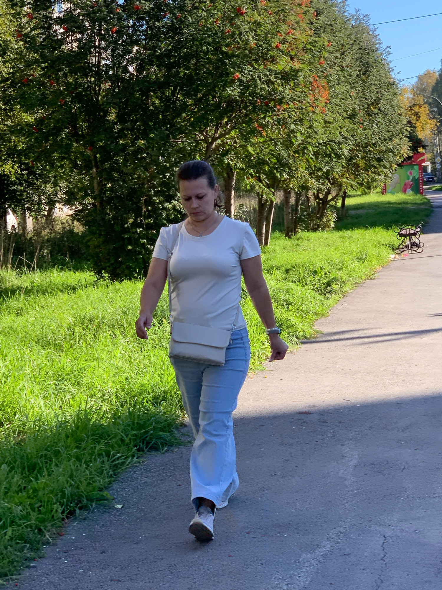 A medium shot shows a woman with dark hair pulled back walking along a paved path lined with lush green grass and trees bearing clusters of red berries. She is wearing a white t-shirt, light blue flared jeans, and white sneakers. A light-colored cross-body bag is slung over her shoulder. Her gaze is directed downward as she walks. The path curves to the right, leading into the distance. To the left of the path, a dense row of trees creates a shaded area. To the right, a black wrought-iron bench sits near the edge of the path, and beyond that, a colorful mural is visible on a building. The scene suggests a park or a tree-lined street in Prokopyevsk, Russia, on a bright, sunny day.