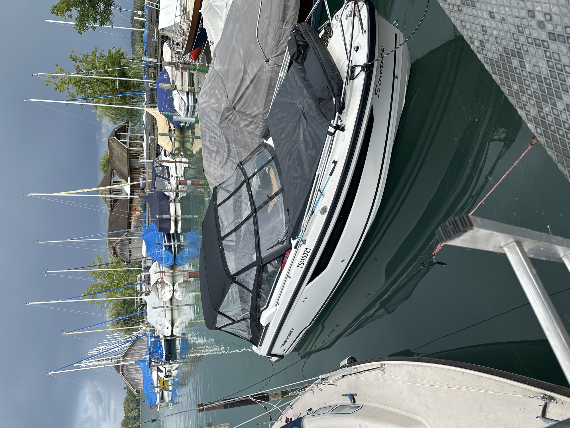 The image depicts a marina on Chiemsee, Germany, with a fleet of sailboats and motorboats docked under a cloudy sky. In the foreground, a white motorboat named "Sunrise" is prominently displayed, its canopy up and a black cover on its bow. Adjacent to it, on the left, is the hull of another boat. In the background, numerous sailboats, many covered with blue and white tarpaulins, line the dock. Traditional wooden boathouses with steep roofs are visible behind the boats, further suggesting the location's setting. The water is calm and reflects the overcast sky and the surrounding vessels. The overall scene conveys a sense of quietude, with the boats at rest in the harbor. There are no people visible in the image.