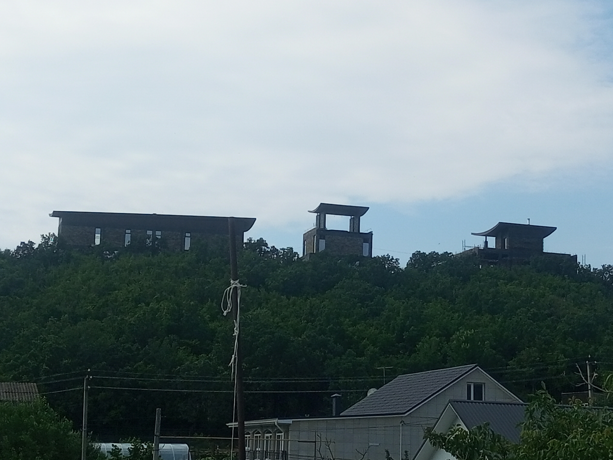 The image depicts a hilly landscape covered in dense green trees. Three modern, multi-story structures with dark, angular roofs are perched on the crests of these hills, overlooking a lower-lying area with residential buildings and utility poles. The structures appear to be constructed from stone or brick, with large windows. In the foreground, a weathered utility pole with a rope tied to it stands prominently. Power lines are visible crisscrossing the scene. The sky is overcast with a light blue hue. The location is specified as Щебетовский поселковый совет, Ukraine.