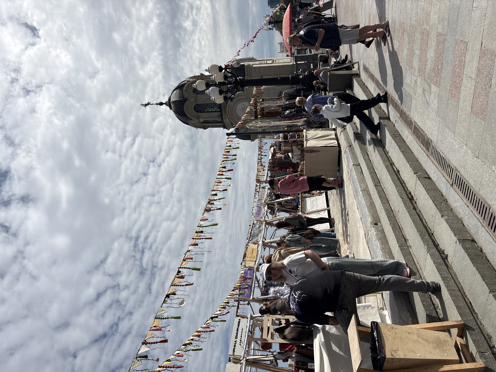 The image captures a scene at the bottom of the stairs in город Ялта, Ukraine. People are browsing vendor stalls set up along the stairs. A building with a dome is visible behind the stalls and colorful decorative banners are strung overhead. The sky is cloudy. People are walking and looking at merchandise. There is a sewer grate running down the staircase. The stairs are constructed of light colored stone.