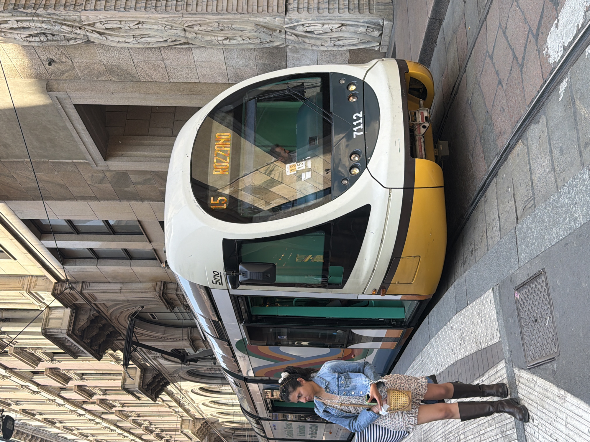 A tram traveling on tram tracks is depicted in a medium shot. The tram is modern, white and yellow, with the number "7112" visible on its front. The tram's digital display reads "15 ROZZANO." A woman stands beside the tram, wearing a denim jacket, a patterned dress, and tall brown boots. She holds a small, woven bag. The tram and woman are positioned on a sidewalk or tram stop. The background reveals a street with ornate, historic buildings lining one side. The location is most likely in Milan, Italy, given the style of the tram and architecture.