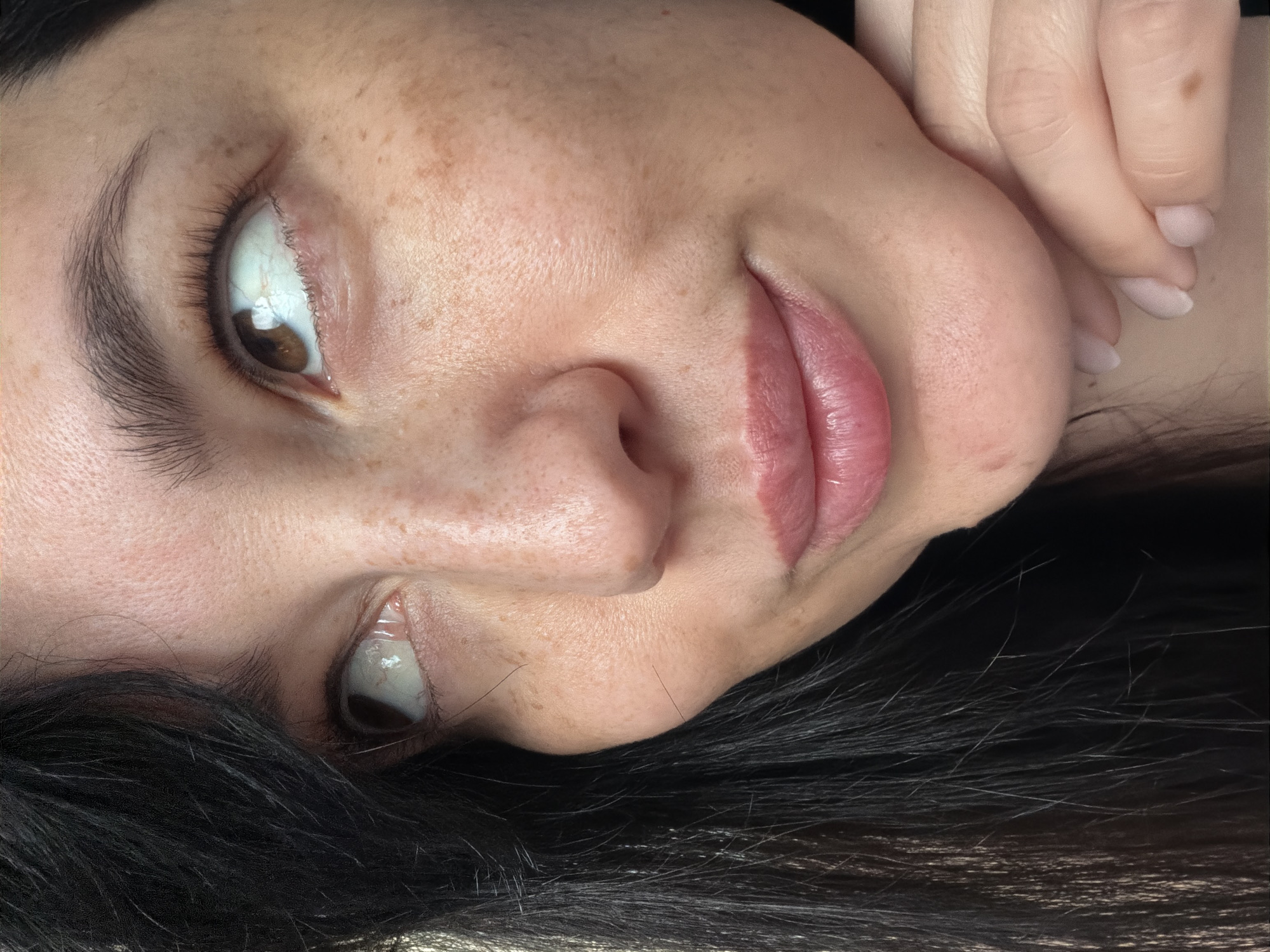 A close-up shot shows the profile of a woman with dark hair and fair skin with freckles. She is looking upwards and to her right. Her left hand is resting on her chin. The image is a portrait and no specific location is visible.