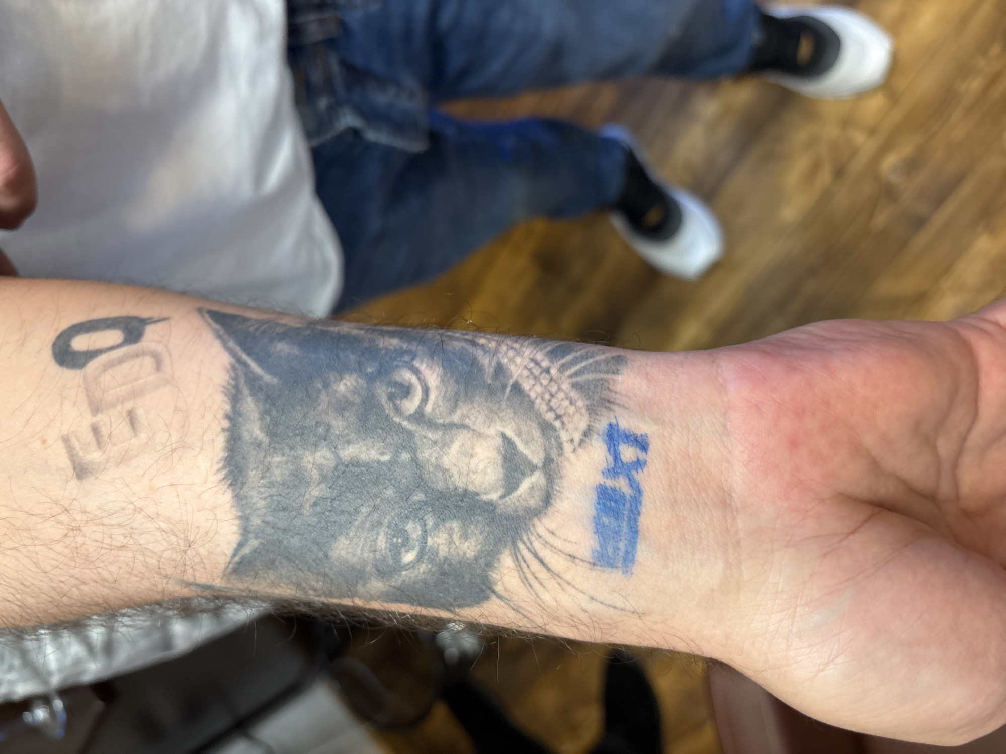 The image shows a person's arm with a tattoo of a cat's face. The tattoo is black and grey, and the cat has large eyes and whiskers. To the left of the cat tattoo, there are some blurred letters or numbers in black. To the right of the cat's face, there is a small, rectangular tattoo in blue ink that appears to have some sort of symbol or writing inside it. The arm is hairy, and the skin looks slightly red in some areas, suggesting it may be a new tattoo or irritated. The background is blurred, but it appears to be an indoor setting with a wooden floor. The person is wearing blue jeans and white shoes, and part of a white shirt is visible. The lighting suggests it is daytime, but there are no specific clues about the time of day or weather. There is no clear indication of emotions. The visible text is "EDD" and a blue symbol or writing.