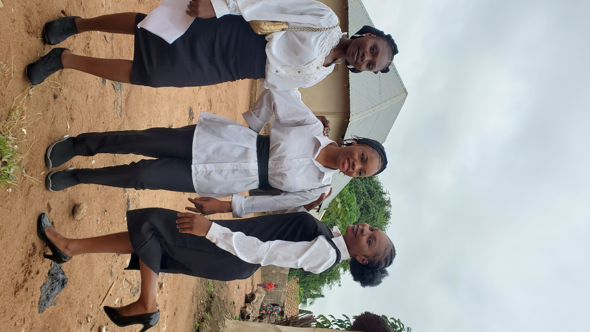 Three young Black women are posing for a photo in an outdoor setting. The woman on the left is wearing a white blouse with intricate gold-colored detailing around the neckline and cuffs, a black knee-length skirt, and black low-heeled shoes. She is holding a white piece of paper in her left hand and has a gold purse slung over her right shoulder. The woman in the center is wearing a white button-up shirt with a black wide belt, black pants, and black slip-on shoes. She has her left arm around the shoulder of the woman on the left. The woman on the right is wearing a white collared shirt with a black sleeveless vest over it and a black skirt that ends just above her knees. She is wearing black high heels and has her right leg bent and raised slightly. She has her left arm around the shoulder of the woman in the center. All three women have their hair styled in braids.
The background suggests a rural or semi-urban environment. A simple house with a corrugated metal roof is visible behind the women. Trees and vegetation are present in the background, as well as a dirt path or road. There are other structures and people in the distance, including what appears to be a small child. The sky is overcast. The overall scene appears to be in a residential area, likely Jalingo, Nigeria, given the architectural style and vegetation.
