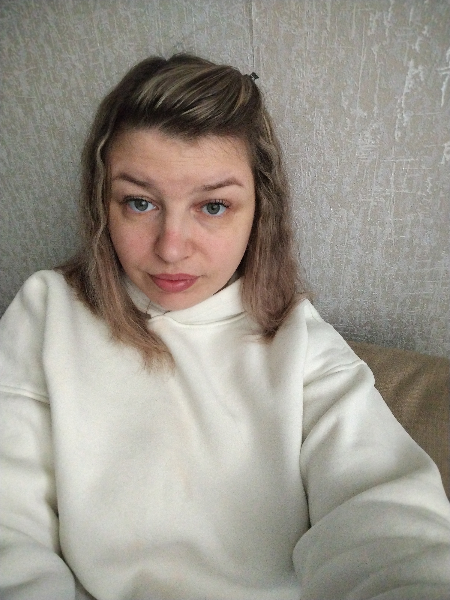 A woman with fair skin, light brown hair with blonde highlights, and blue eyes is wearing a white hoodie. Her hair is partially pulled back with a clip. She is indoors with a textured grey wall behind her and a beige sofa beneath. The photo appears to be a selfie. There is no indication of the location being Шебунино, Russia.