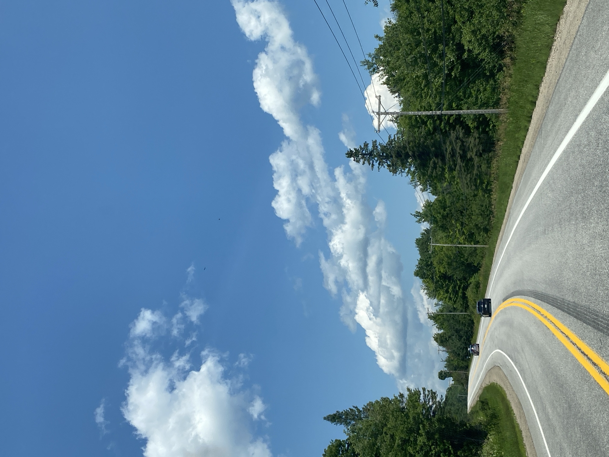 The image depicts a two-lane asphalt road curving through a rural landscape under a bright blue sky with scattered white clouds. The road is marked with double yellow lines in the center and white lines on the edges. Several dark-colored cars are visible on the road, with one in the foreground and two further ahead. The roadside is lined with lush green trees and vegetation. Telephone poles with wires are interspersed along the side of the road. There are no visible buildings or signs of a city. The scene appears to be a sunny day in a natural environment, not within the city of Ripon, Canada, as there are no urban features present.