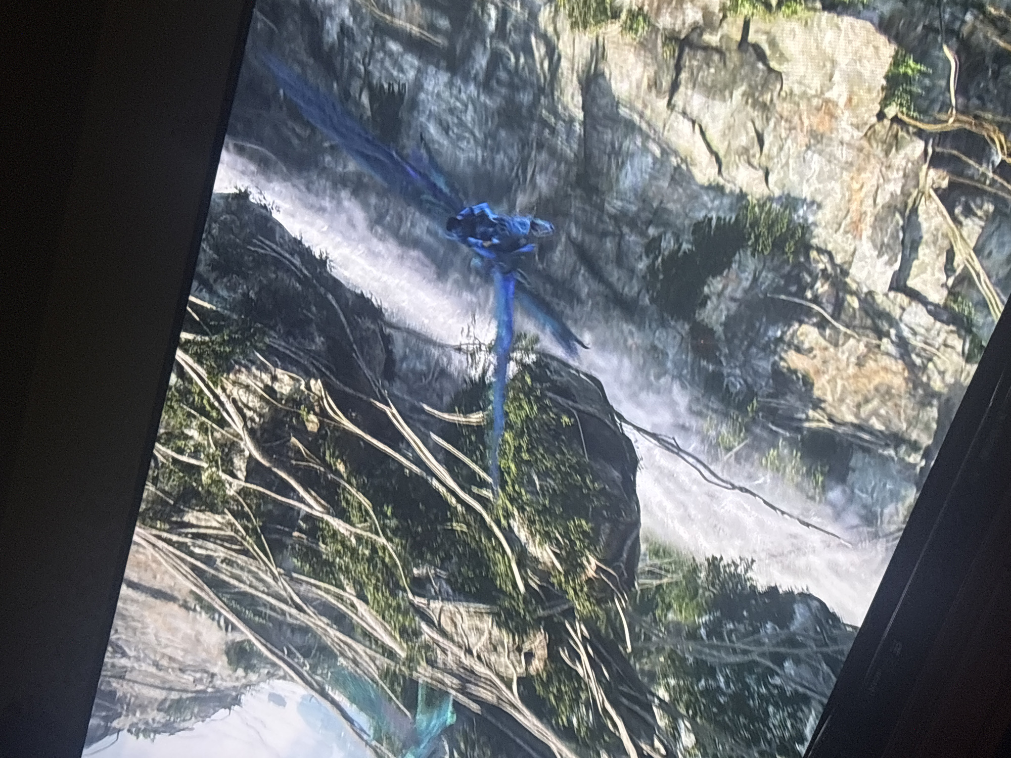 A blue flying creature with large wings soars past a waterfall against a rocky cliff face covered in vegetation. The scene is viewed on a television screen. The location is not discernible as San Pablo del Monte, Mexico.