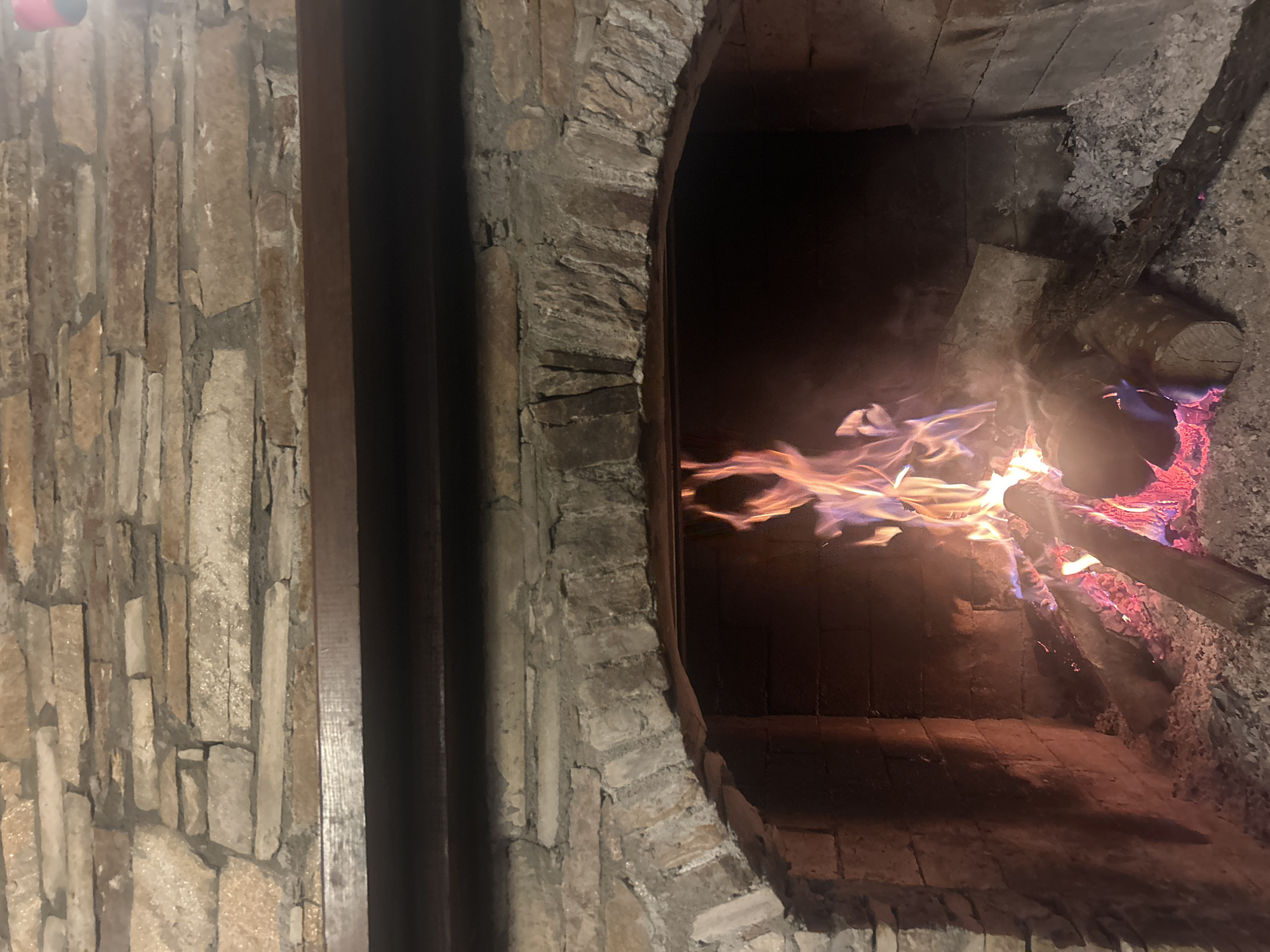 The image shows the interior of a brick oven. Flames are visible, consuming logs inside the oven. The oven's walls and arched entrance are constructed of irregular, tan-colored stones. A section of a wall, also built of similar stones, is on the left side of the frame. The oven is possibly located in Dolen, Bulgaria.