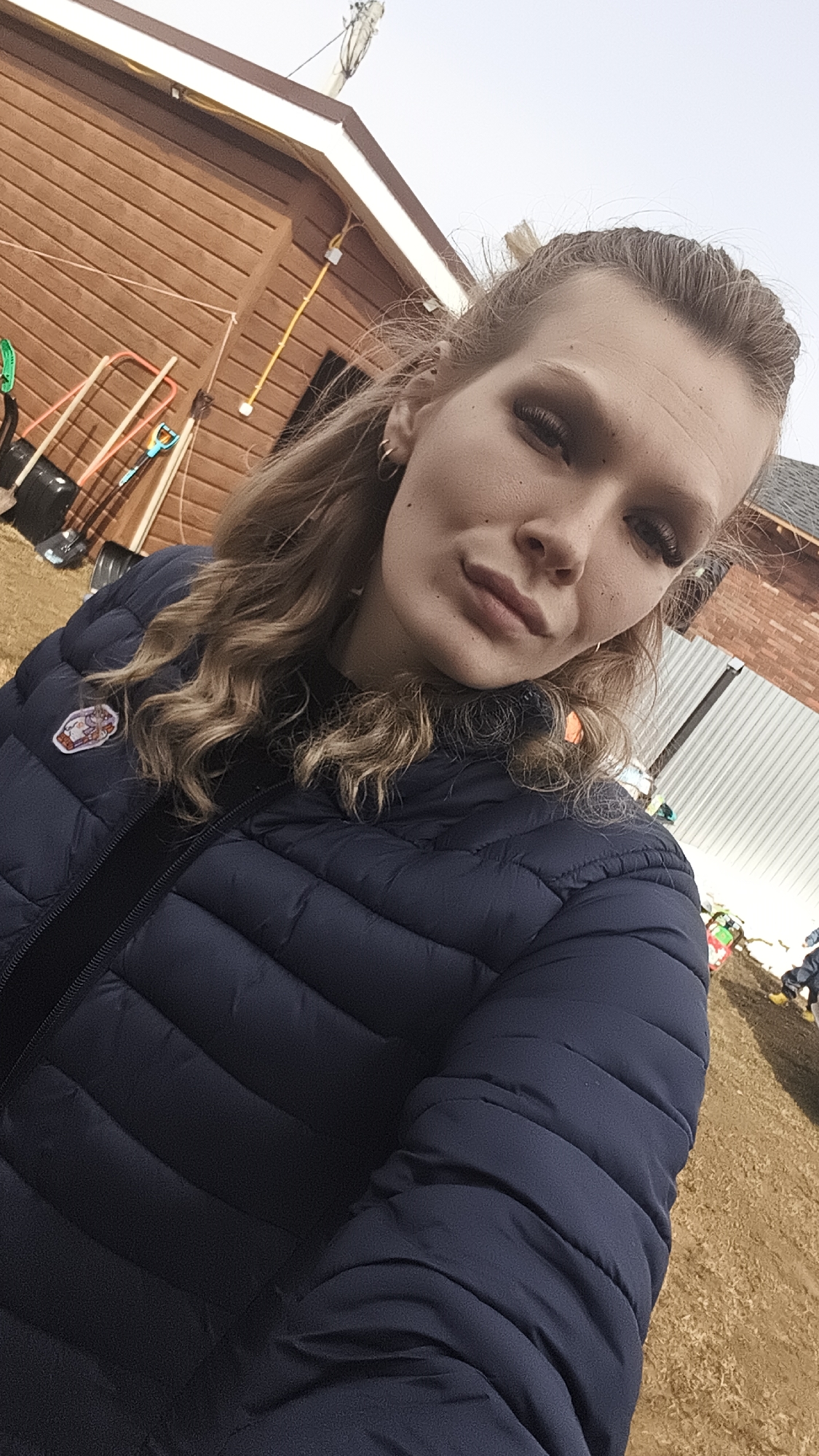 The image is a selfie of a young, fair-skinned individual with light brown hair. They are wearing a dark blue, quilted puffer jacket. The background suggests an outdoor setting with a brown wooden building and various gardening tools visible. There's also a patch of dry, grassy ground visible. The photograph was taken during the daytime, likely in Русский Вожой, Russia, as specified in the request.