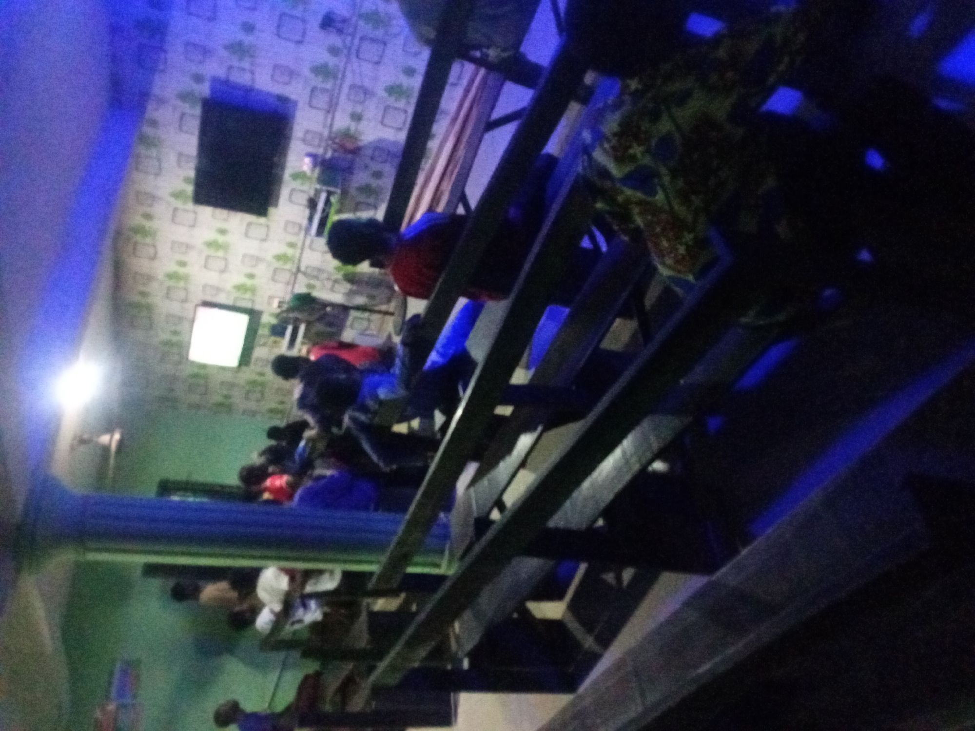 The image depicts an interior space in an urban location. The scene features multiple individuals sitting at tables and appearing to be focused on screens, likely in a gaming or internet cafe. The setting seems to be a multi-level establishment, as suggested by the structural elements that could be part of a balcony or upper seating area. The walls are adorned with patterned wallpaper. The lighting is dim, with a noticeable blue hue that adds to the ambiance of the place. The location is in the city of Unknown, Unknown.