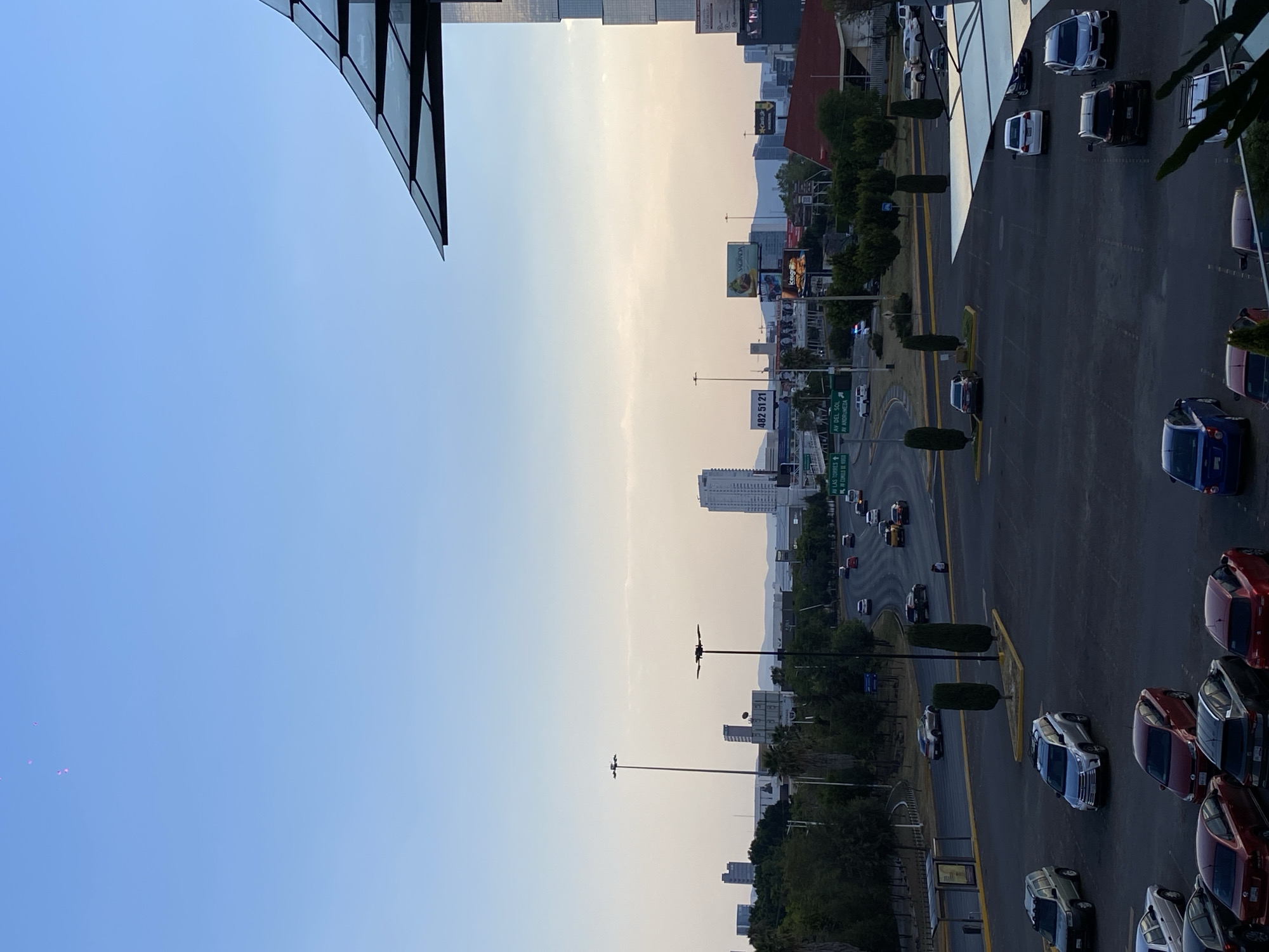 The image presents an elevated view of a city street in San Bernardino Tlaxcalancingo, Mexico. The scene is dominated by an asphalt road with multiple lanes, delineated by yellow lines, with cars parked or traveling along it. A landscaped median separates the lanes. The surroundings include vegetation, buildings of various heights, and billboard advertising. Streetlights are visible along the roadway. The sky, transitioning from light blue to pale yellow, suggests either sunrise or sunset. Part of a modern building with glass panels is visible in the upper-left corner. Street signs indicate "AV. DEL SOL/AV ANDROMEDA" and "LAS TORRES/KAF CUNILA DE VASO".