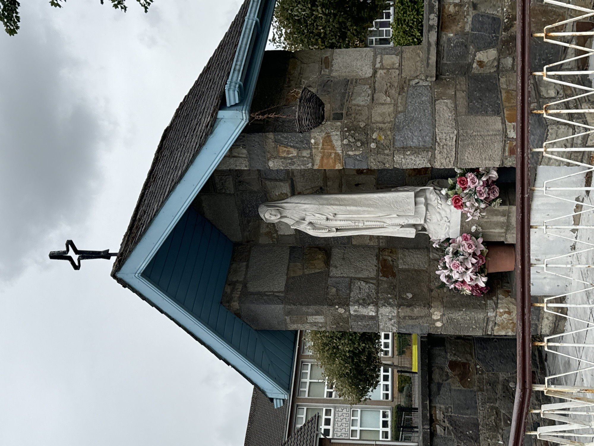 The image depicts a small, open-air shrine constructed with rough-hewn stone. A white statue of the Virgin Mary, holding rosary beads, stands centrally within the shrine. Two pots of artificial flowers, one with pink and white roses and another with predominantly pink roses, are placed at the base of the statue. Above the shrine, a dark, stylized cross is mounted on the roof. The roof itself is shingled and edged with light blue trim. A black woven basket hangs from a chain on the right side of the shrine's overhang. In the background, a multi-story building with numerous windows is visible, along with trees and a cloudy sky. The foreground features a white, metal railing with a dark red top. This shrine is located at the entrance to the Mercy Primary School on Trinity Without in the city of Waterford, Ireland.