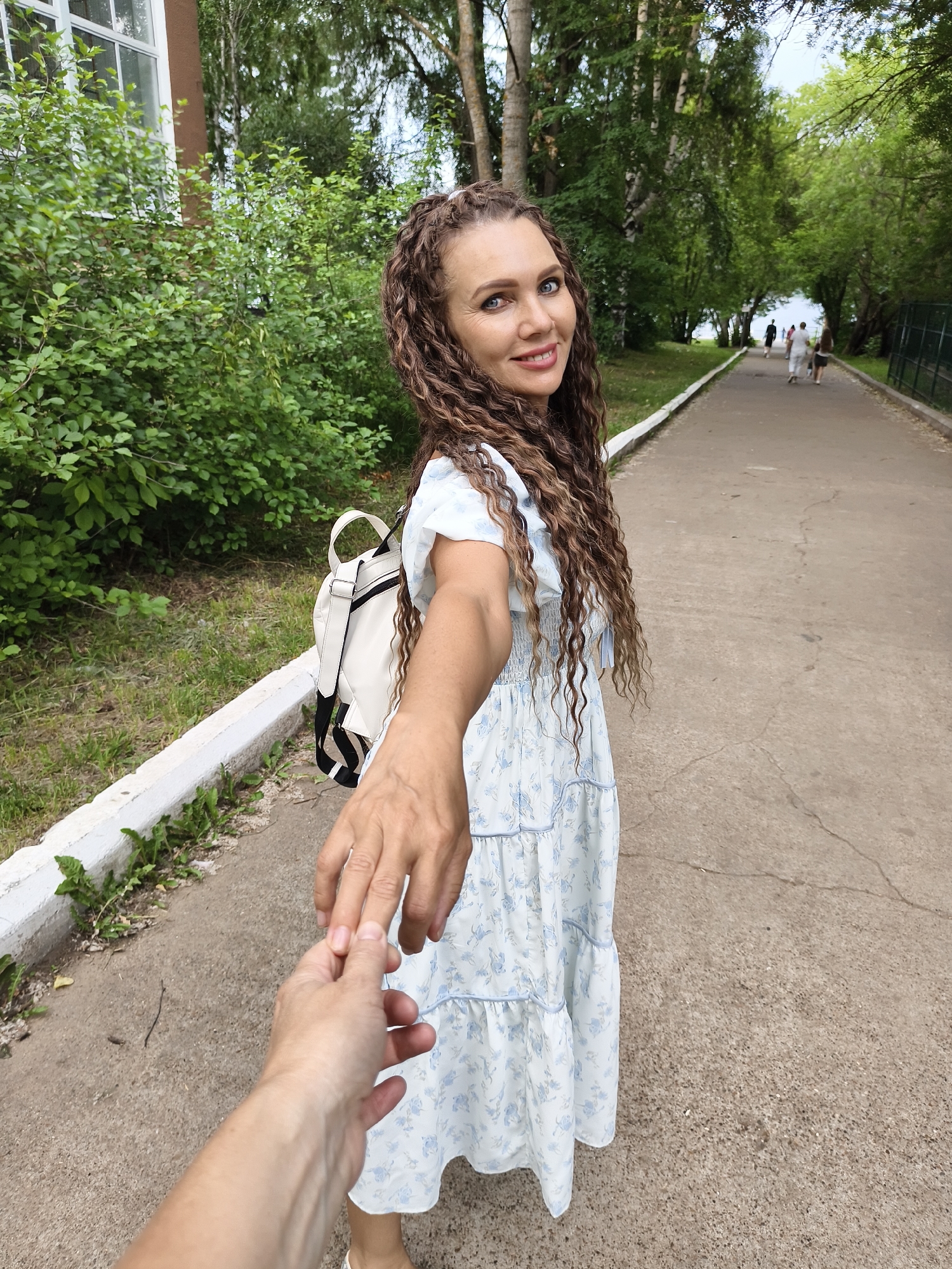 A woman with long, curly brown hair, wearing a light blue and white floral dress and a white backpack, holds out her hand to the viewer as if inviting them to follow. She is smiling and looking at the camera. The scene is outdoors, on a paved pathway lined with greenery and trees. In the background, other people are walking along the path, and a body of water is visible in the distance. The time of day appears to be daytime, with overcast skies suggesting a mild or warm weather. The setting is likely a park or recreational area in Chaikovsky, Russia, given the context. There is no visible text in the image.