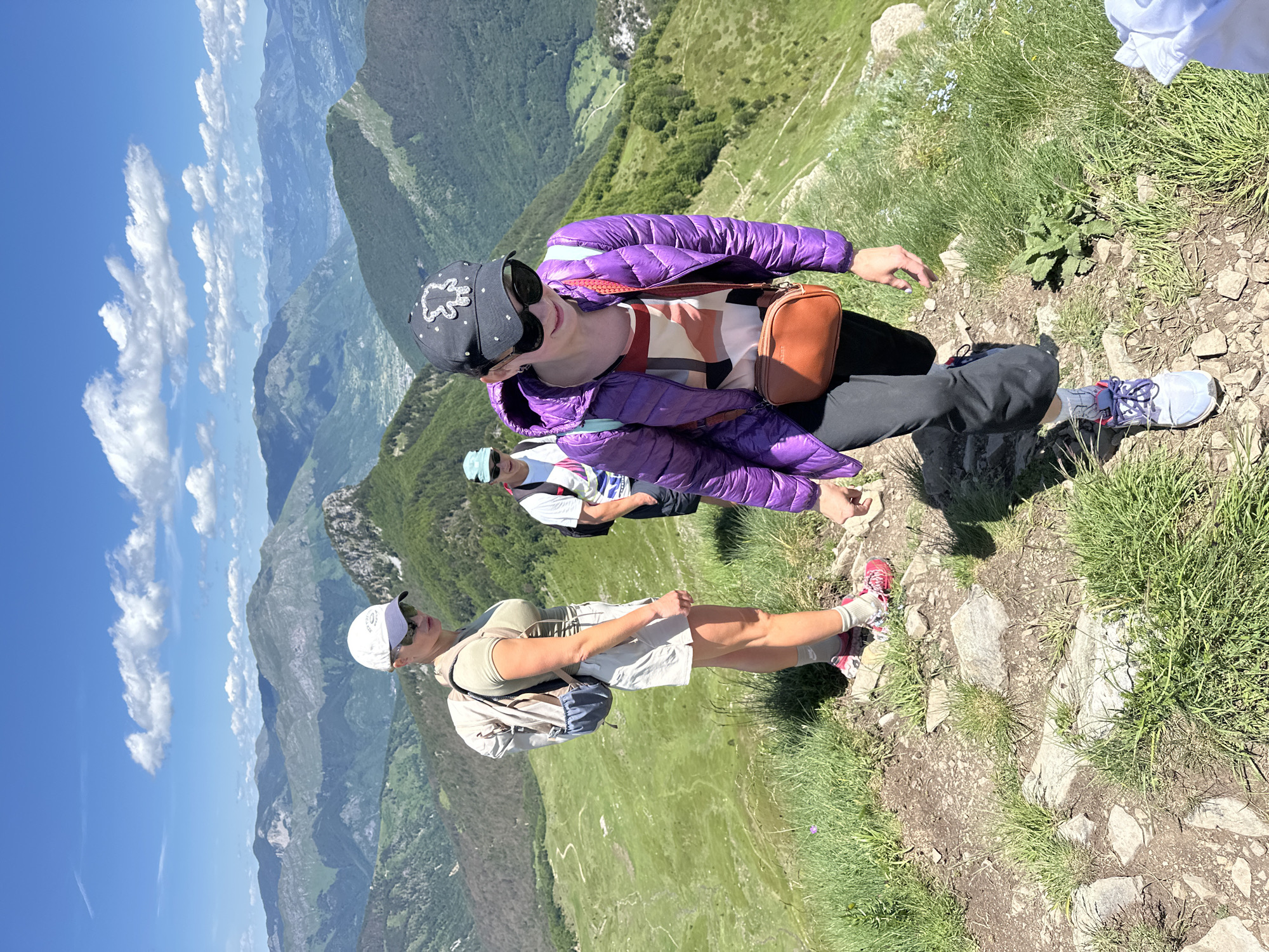 In the city of Dolja, Montenegro, three individuals are hiking on a mountain trail under a clear blue sky with scattered white clouds. The person in the foreground, wearing a purple puffer jacket, dark pants, and a black baseball cap with a teddy bear design, is walking downhill. To their left and slightly behind, a person in a light green t-shirt and white shorts, wearing a white baseball cap and sunglasses, is walking uphill, carrying a backpack. Further back and to the left, a third person wearing a white t-shirt and a blue baseball cap is also hiking uphill. The terrain is rocky with patches of green grass and vegetation. The background reveals a vast mountainous landscape with lush green slopes and distant hazy mountains.