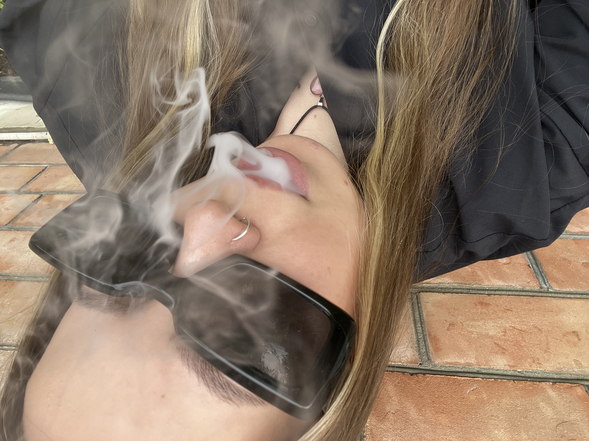 A young woman lies on a brick surface. Her head is oriented towards the lower left corner of the frame. She wears dark sunglasses. Smoke emanates from her mouth. She has blonde hair. The location is Novopokrovskaya, Russia.