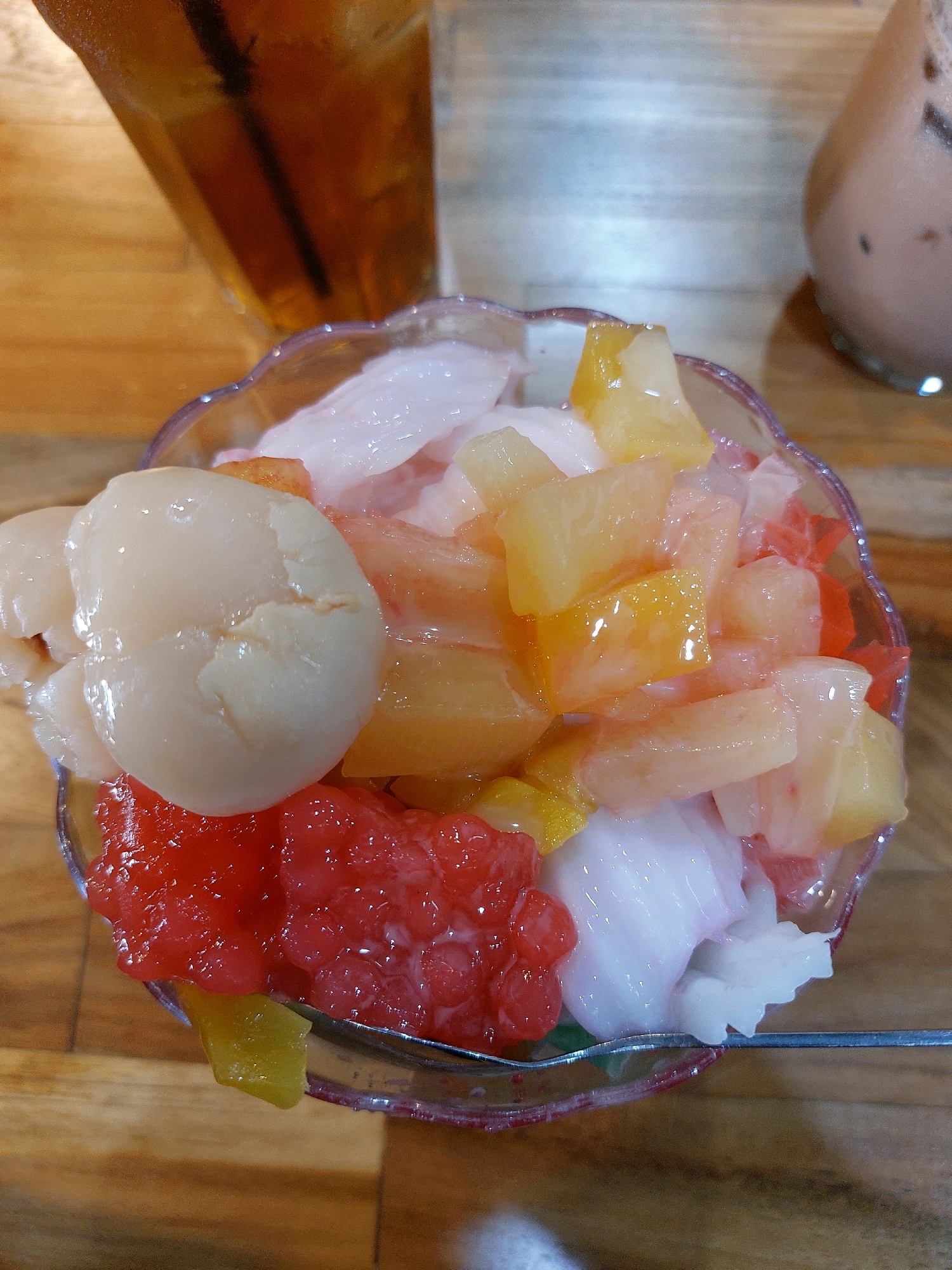 A bowl of fruit cocktail sits on a wooden table, with a glass of iced tea in the background. The fruit cocktail contains lychee, jellied watermelon, pineapple, and possibly other fruits, all mixed with a creamy white liquid. The bowl has a decorative rim. The setting appears to be an eatery in Tarakan, Indonesia.