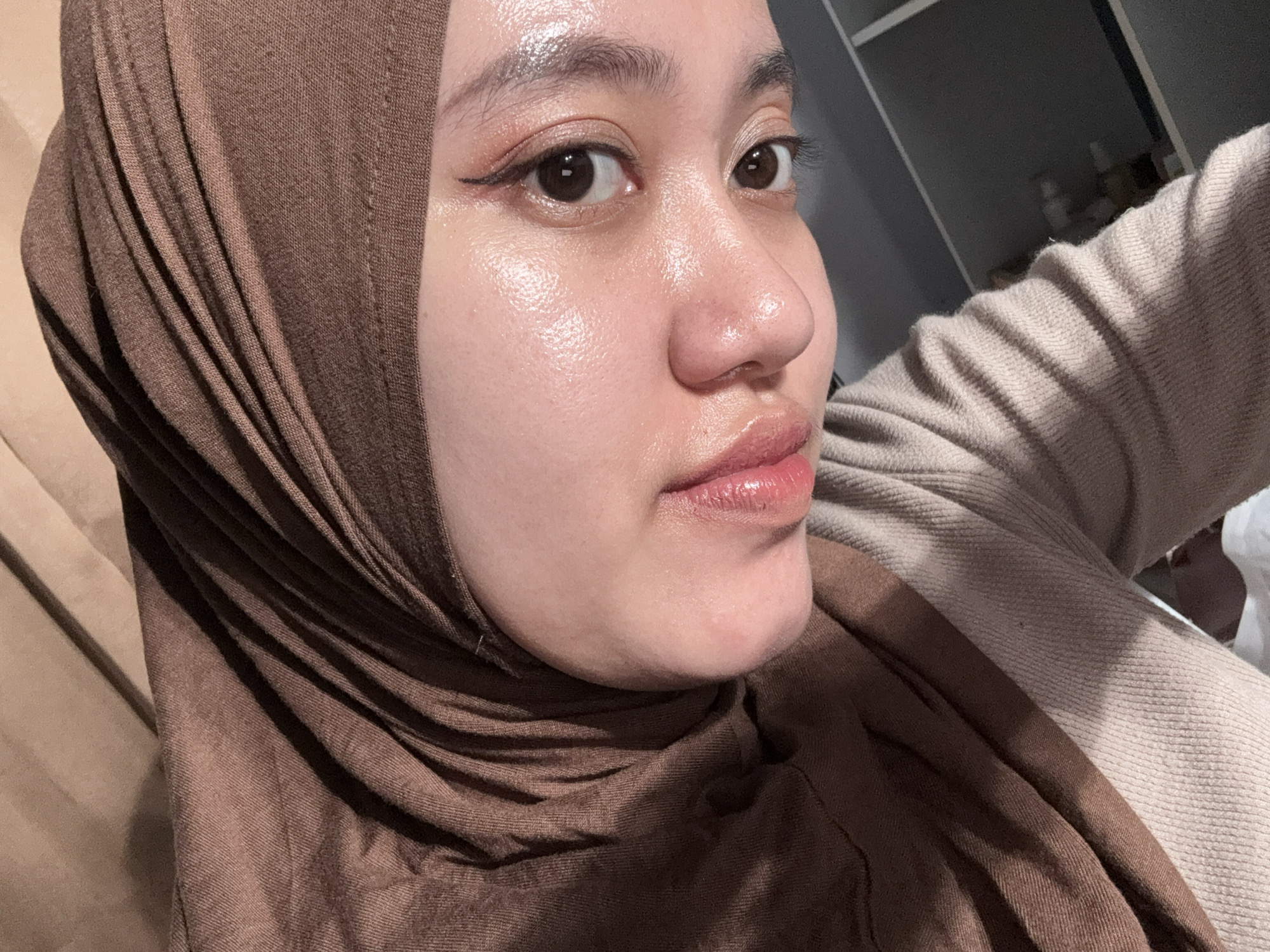 A close-up photograph depicts a person wearing a brown hijab. The individual has fair skin, brown eyes, and is wearing subtle makeup, including eyeliner. The person is wearing a beige colored sweater. The background is blurry, but includes shelves and objects that suggest an indoor setting. There is nothing in the image to suggest a location in Gadingrejo, Indonesia.