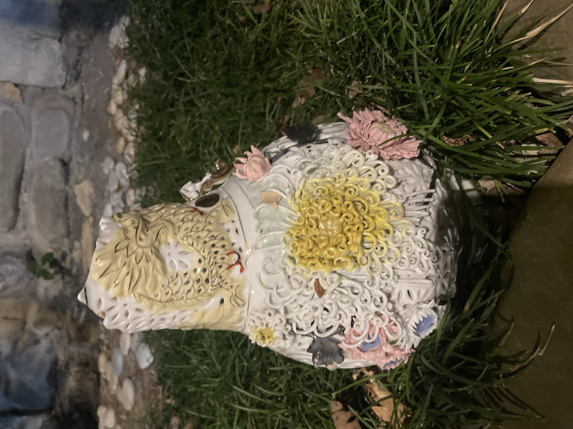 A white porcelain vase with intricate detailing is placed on green grass. The vase features a dragon motif on its upper portion, rendered in yellow and gold. The lower part of the vase is adorned with a large, textured yellow flower and several smaller pink and light blue flowers, as well as delicate white filigree work. The background consists of a stone wall and small pebbles. The lighting suggests it is evening or nighttime.