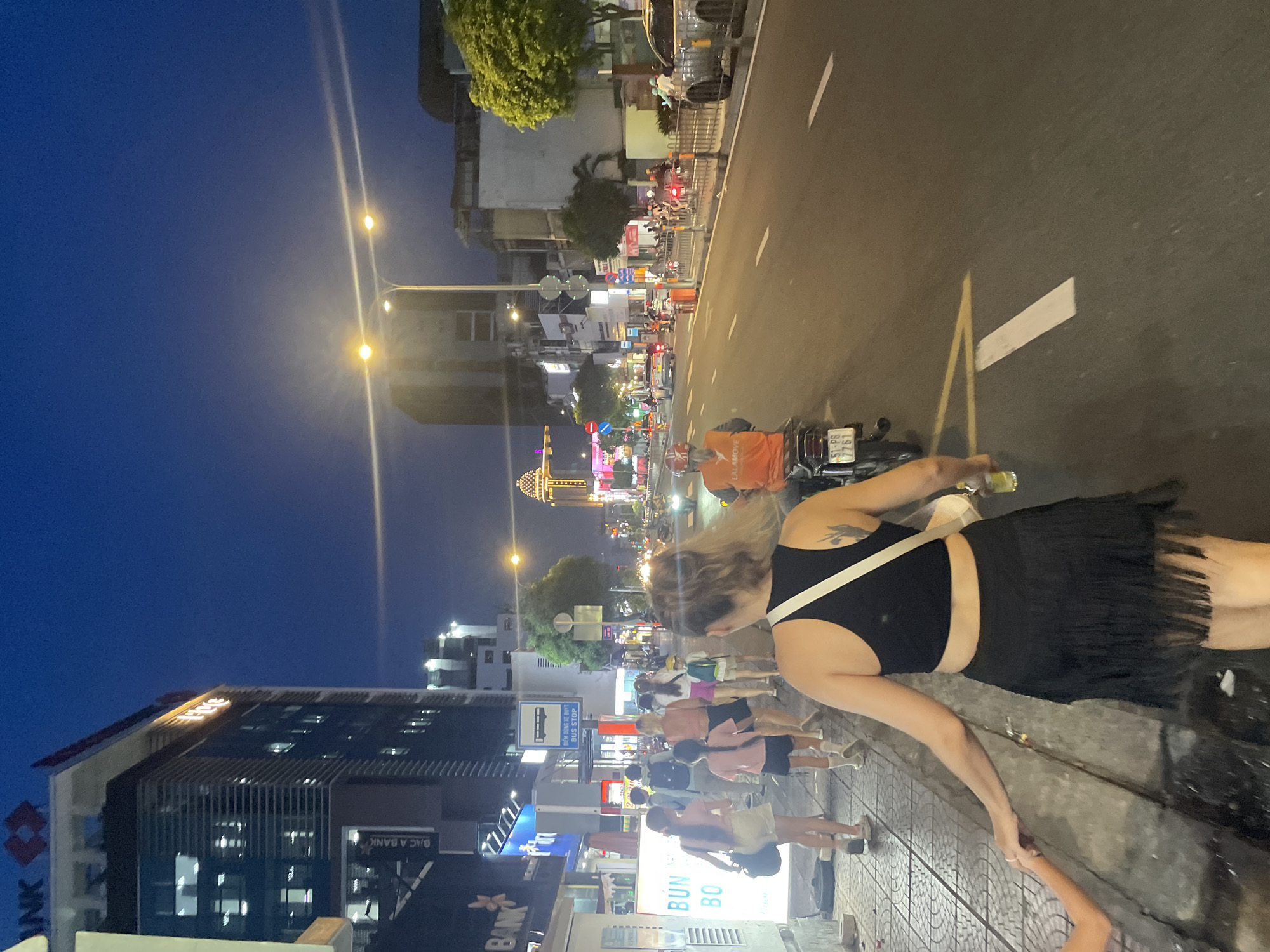 The image captures a lively street scene at dusk in Thủ Đức, Vietnam. A woman in a black top and fringed skirt is the primary subject, her arm extended as if reaching out or walking. Behind her, a street bustles with activity. Several people are walking on the sidewalk, many appearing to be young women in casual attire, carrying bags. A motorcycle with an orange LALAMOVE bag is parked nearby, with a license plate visible (51-P8 7761).
The setting is a city street lined with buildings, some featuring illuminated signs, suggesting commercial activity. Streetlights cast a warm glow on the road and sidewalk, creating a vibrant evening atmosphere. Signs with Vietnamese text are visible, including "ĐIỂM DÙNG XE BUYT BUS STOP" and "BUN BO," indicating a bus stop and likely a food vendor. The buildings also display logos for "BAC A BANK" and "ANK," identifying financial institutions.
The time of day is evident from the deep blue twilight sky and the artificial lighting illuminating the street and buildings. The overall impression is one of a dynamic urban environment coming alive as night falls, with people engaged in everyday activities like shopping, commuting, or dining. The image evokes a sense of urban energy and cultural vibrancy.