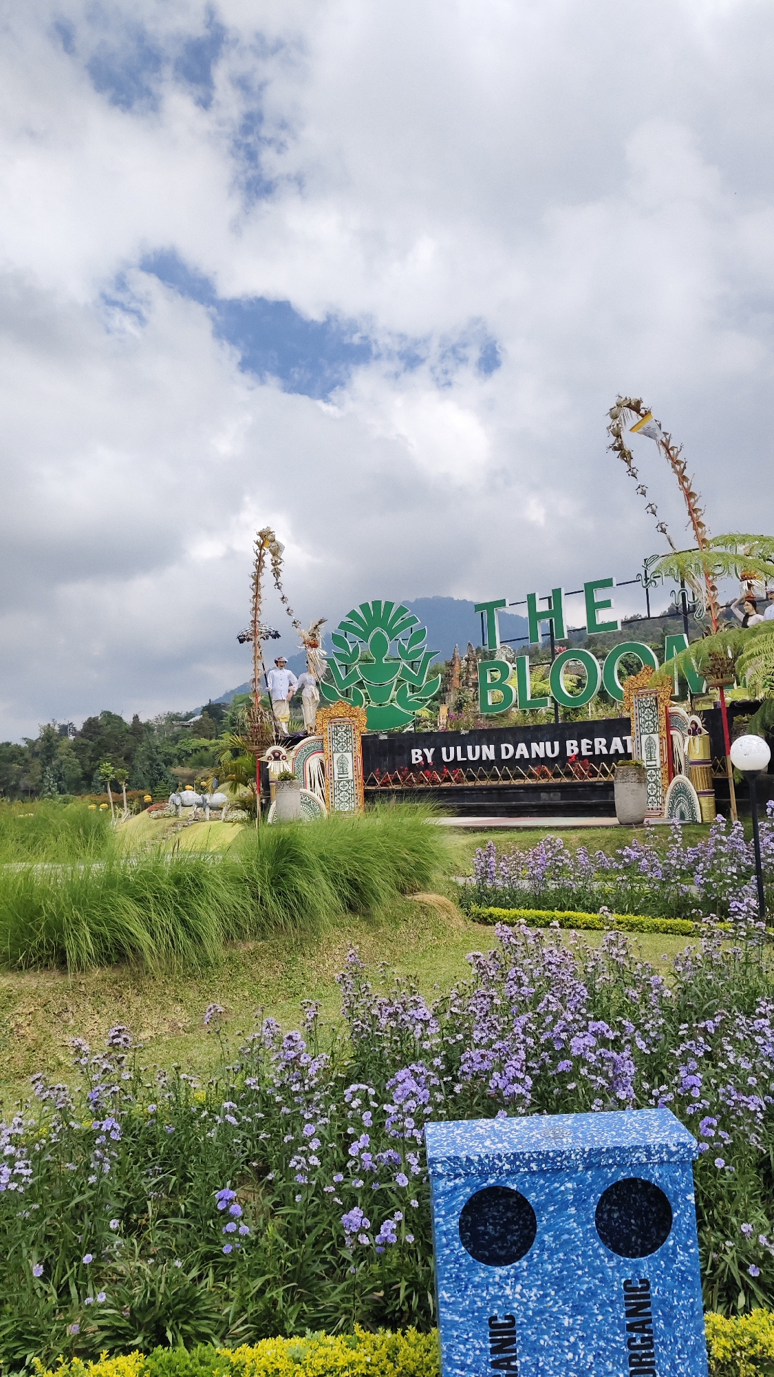 This is a vertical, eye-level, medium shot of a sign that reads "THE BLOOM" with a logo above it. The sign is located in front of a lush green landscape with flowers. The location is identified as Baturiti, Indonesia. In the foreground, there are purple flowers and a blue recycling bin. Behind the sign, there is a mountain and trees. The sky is cloudy. The scene appears to be a tourist attraction.