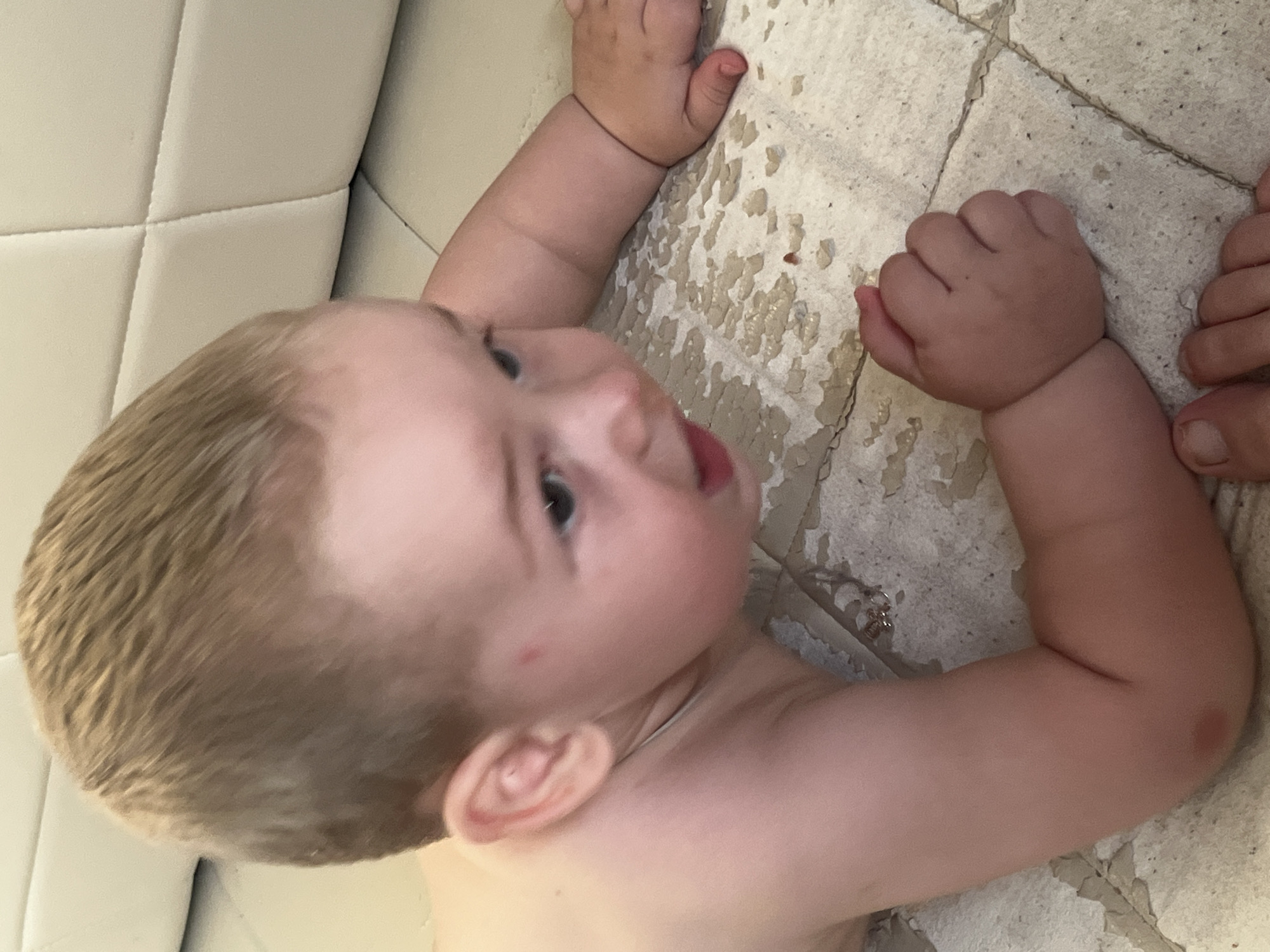 A blond-haired baby is crawling on a tiled floor, which appears wet and possibly peeling in some areas. The baby is wearing no clothes and has a small red mark on its cheek. In the background, a light-colored, cushioned surface, possibly a couch, is visible. The scene suggests an indoor domestic environment. The exact location within Chelyabinsk, Russia, cannot be determined from the visual information provided.