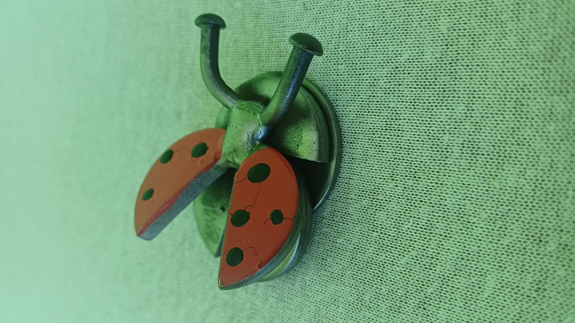 A close-up, high-angle shot shows a decorative metal ladybug ornament resting on a textured, light green fabric surface. The ladybug has a metallic green body and two red wings with black polka dots. The wings are slightly open, revealing the metallic underside. The ornament is positioned in the center of the frame, with the fabric filling the rest of the image. The lighting is soft and diffused, casting no harsh shadows. The location is not discernible from the image.