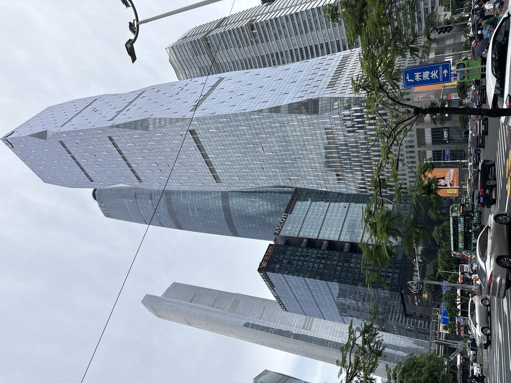 The image captures a street-level view of a bustling urban environment in Tianhe District, China, dominated by towering modern skyscrapers. In the foreground, a multi-lane road is filled with various vehicles including cars, buses, and motorcycles. Pedestrians are visible on the sidewalks and crossing the street. The buildings are a mix of glass-fronted skyscrapers with distinct architectural designs, some featuring intricate geometric patterns and others with more conventional rectilinear forms. One prominent building displays signage for "ASCOTT" and "YUEXIU GROUP". A green double-decker bus with an advertisement on its side is a notable element in the traffic flow. A blue directional sign with Chinese characters indicating "CUSTOMS GUANGZHOU" and an arrow is also visible. Lush green trees line the sidewalks, providing a contrast to the concrete and glass structures. The sky is overcast, suggesting a cloudy day. The overall scene depicts a dynamic city center with significant commercial and transportation activity.