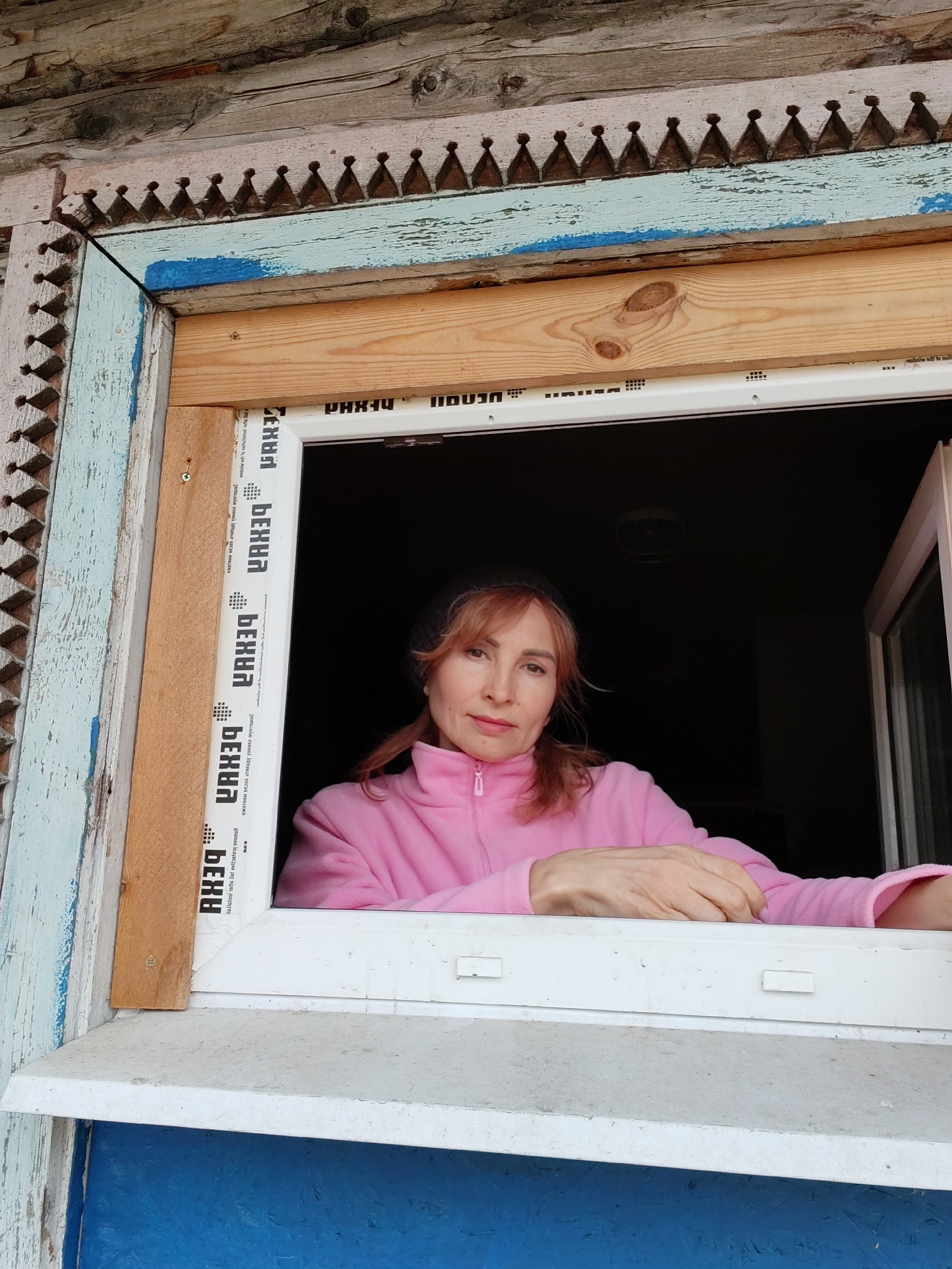 A middle-aged woman with auburn hair peers out of a window. She is wearing a pink fleece jacket. The window is framed by a weathered wooden structure with decorative carvings and peeling blue paint. The window itself is a modern PVC type, featuring the brand "REXAU" (written in reverse) printed on the frame. The interior behind the woman is dark, suggesting nighttime or a dimly lit room. The scene is likely a private residence in Chaikovsky, Russia, based on the architectural style and the presence of the "REXAU" brand.