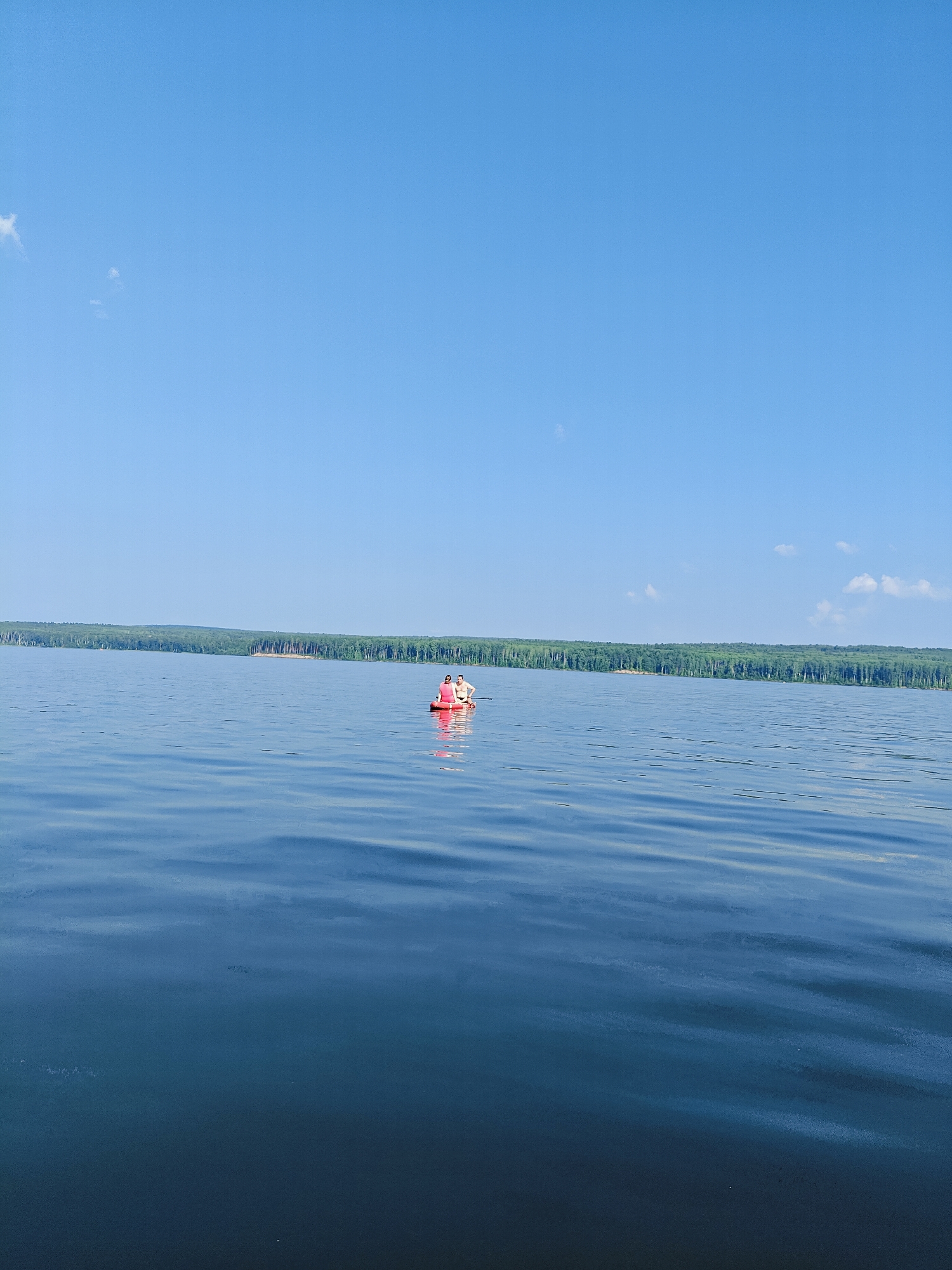 Two people are in a small red raft on a large body of water. They are facing away from the camera, and the person in the front is wearing a pink shirt. The water is calm with slight ripples. In the distance, a dense line of green trees forms the shoreline, suggesting a natural setting. The sky is a clear, bright blue with a few small, wispy clouds. This scene is likely on the Kama River in Chaikovsky, Russia, given the description of the landscape which matches the appearance of the river within the city.